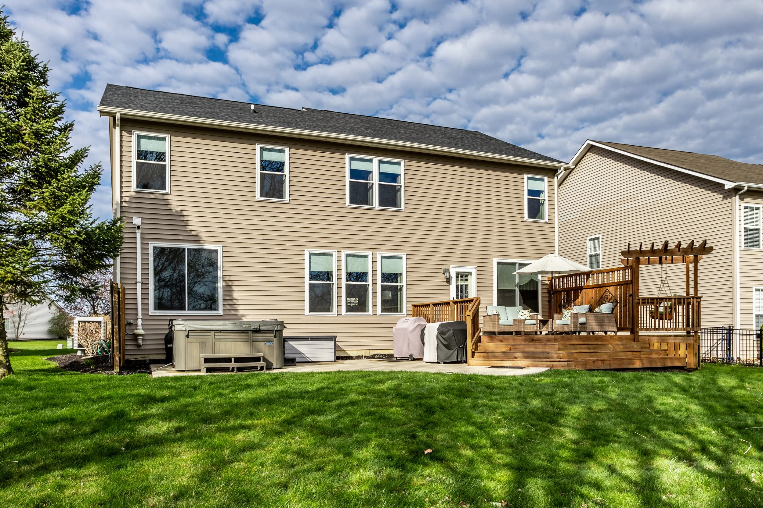 Modern suburban house with beige siding, large windows, and a wooden deck featuring outdoor furniture and an umbrella. The backyard has green grass and is bordered by neighboring houses. A covered grill and hot tub are also visible.