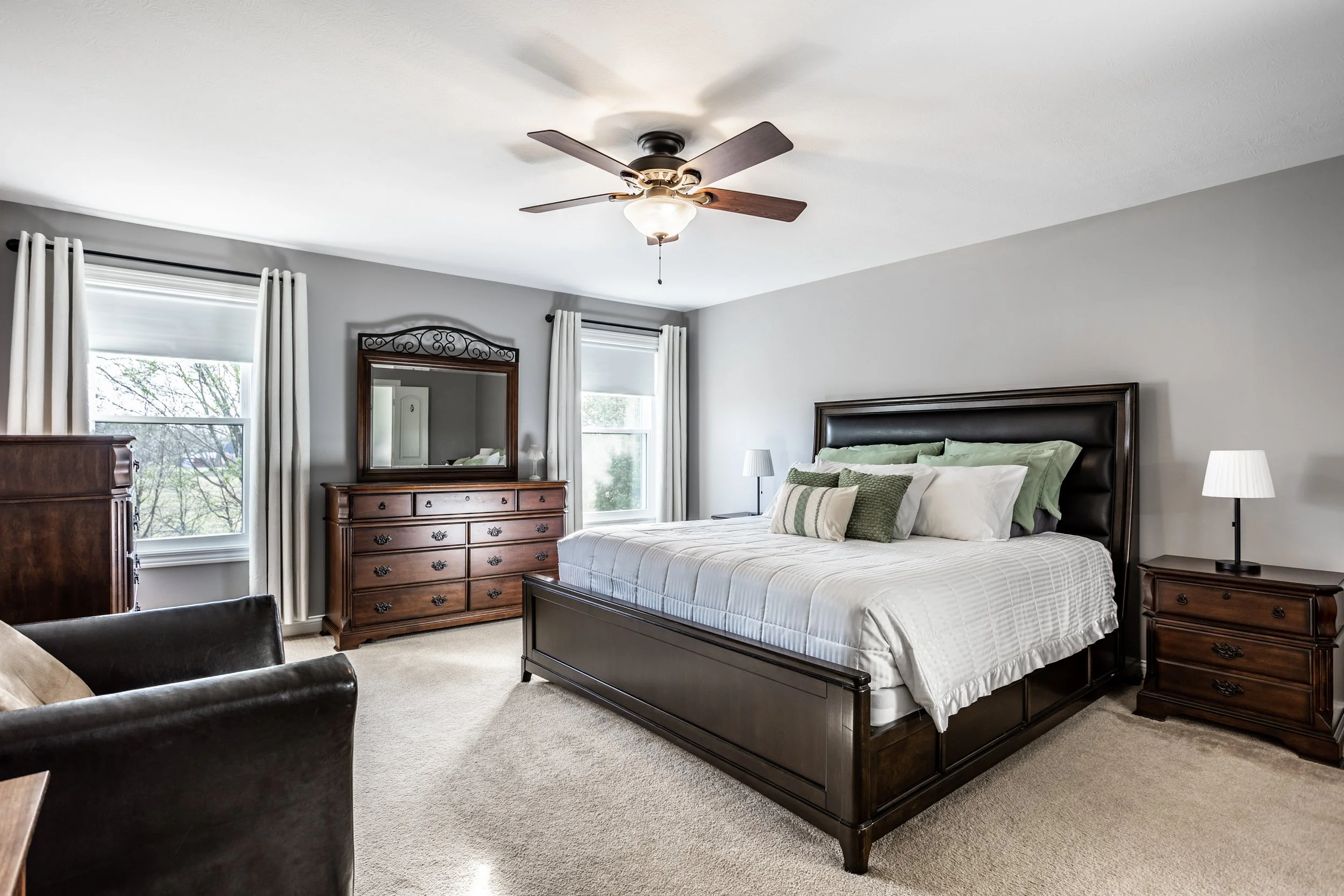 Modern bedroom with a large bed, dark wood furniture, ceiling fan, and two windows with white drapes.