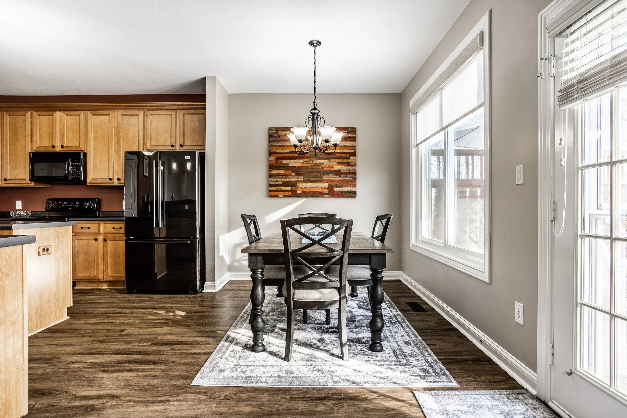 Modern kitchen and dining area with wooden cabinets, black appliances, a dining table with chairs, a chandelier, and large windows with natural light.