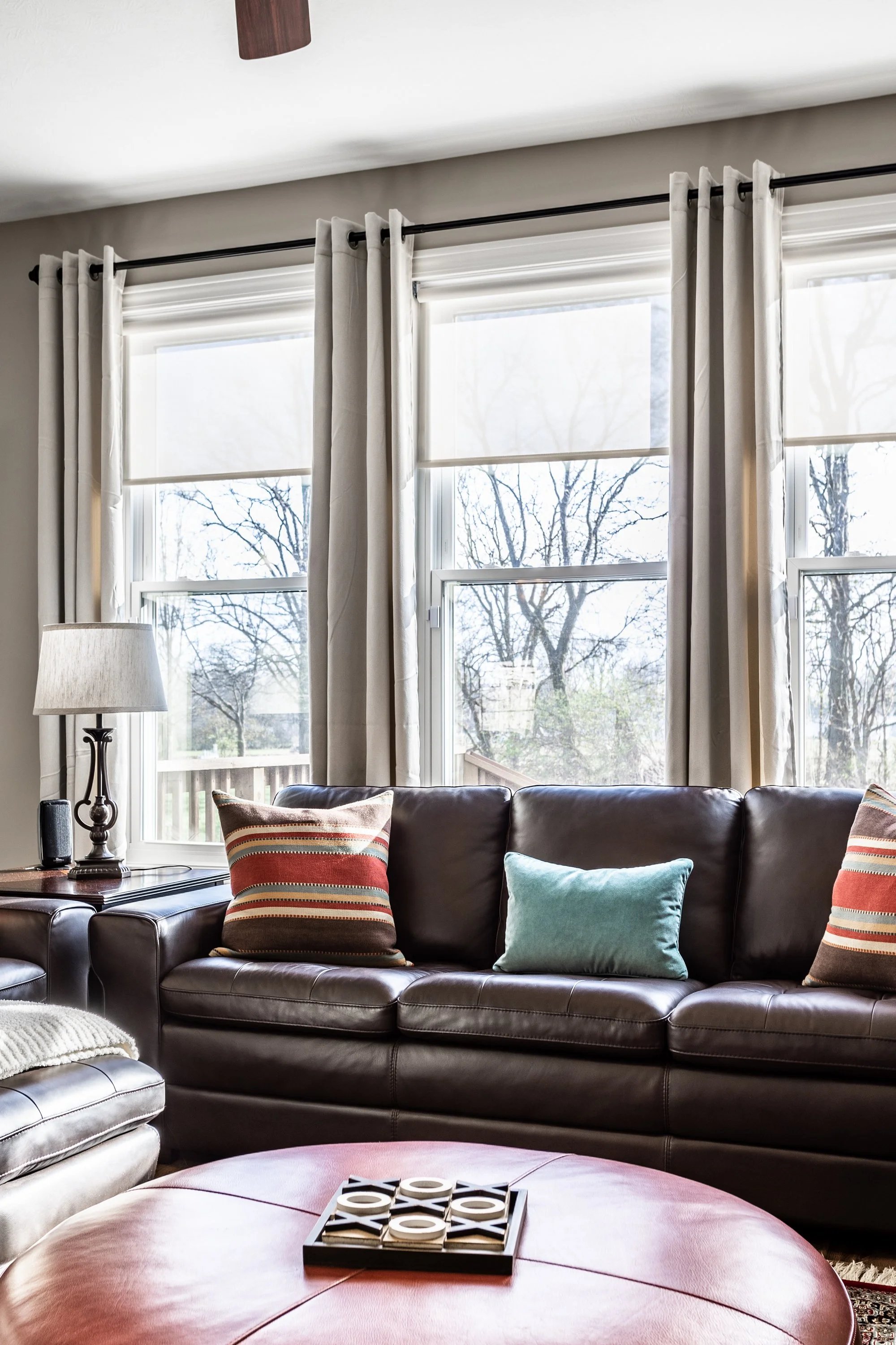 Living room with brown leather sofa, colorful throw pillows, round leather ottoman, lamp, and large windows with curtains.