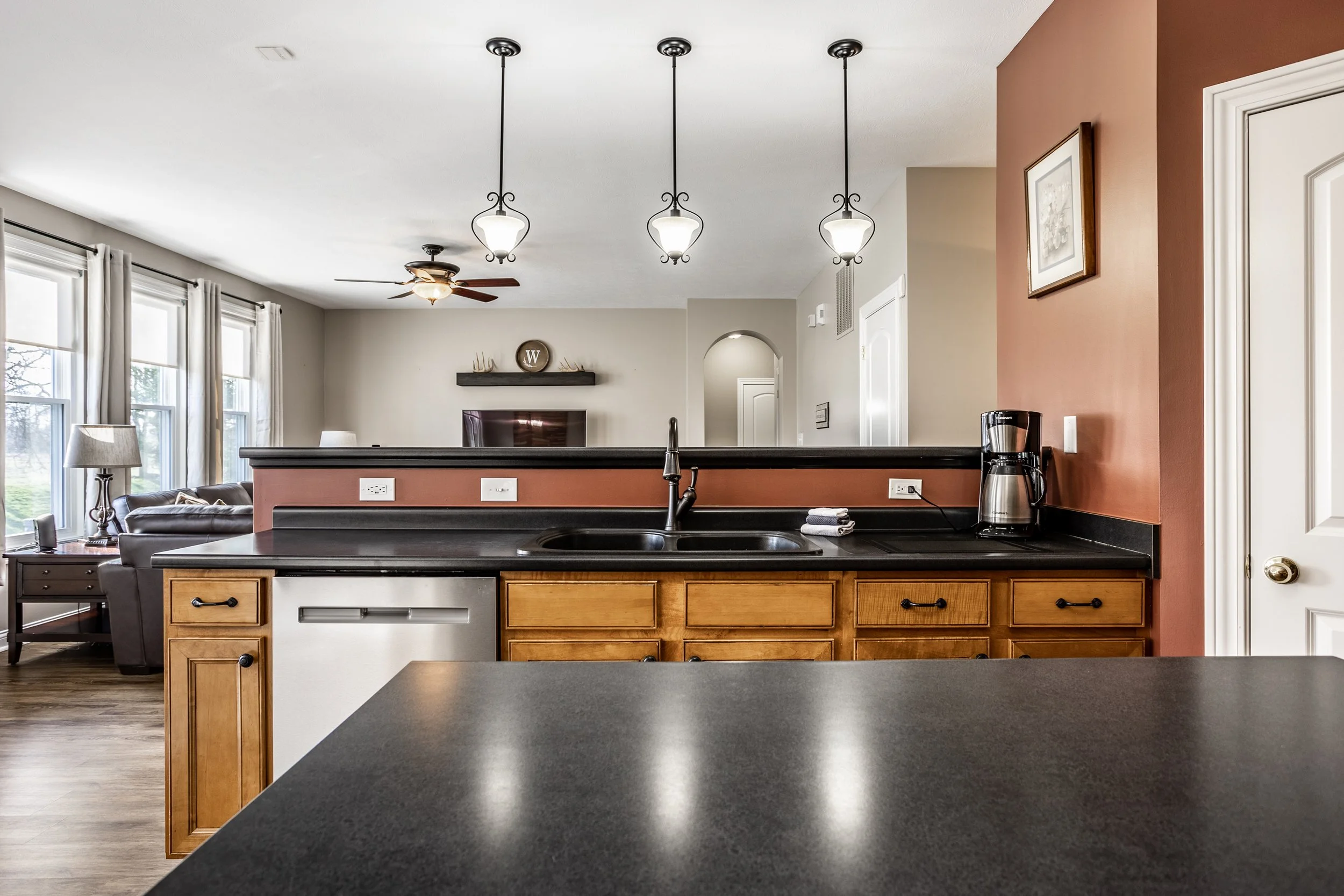 Modern kitchen and living room with a black countertop, stainless steel coffee maker, wooden cabinets, pendant lights, ceiling fan, and large windows.