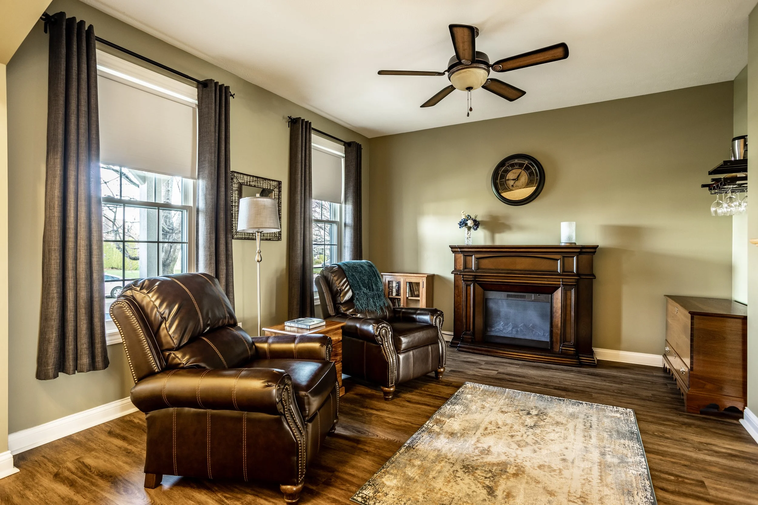 Cozy living room with two leather armchairs, wooden floor, ceiling fan, wall clock, electric fireplace, and decorative items.