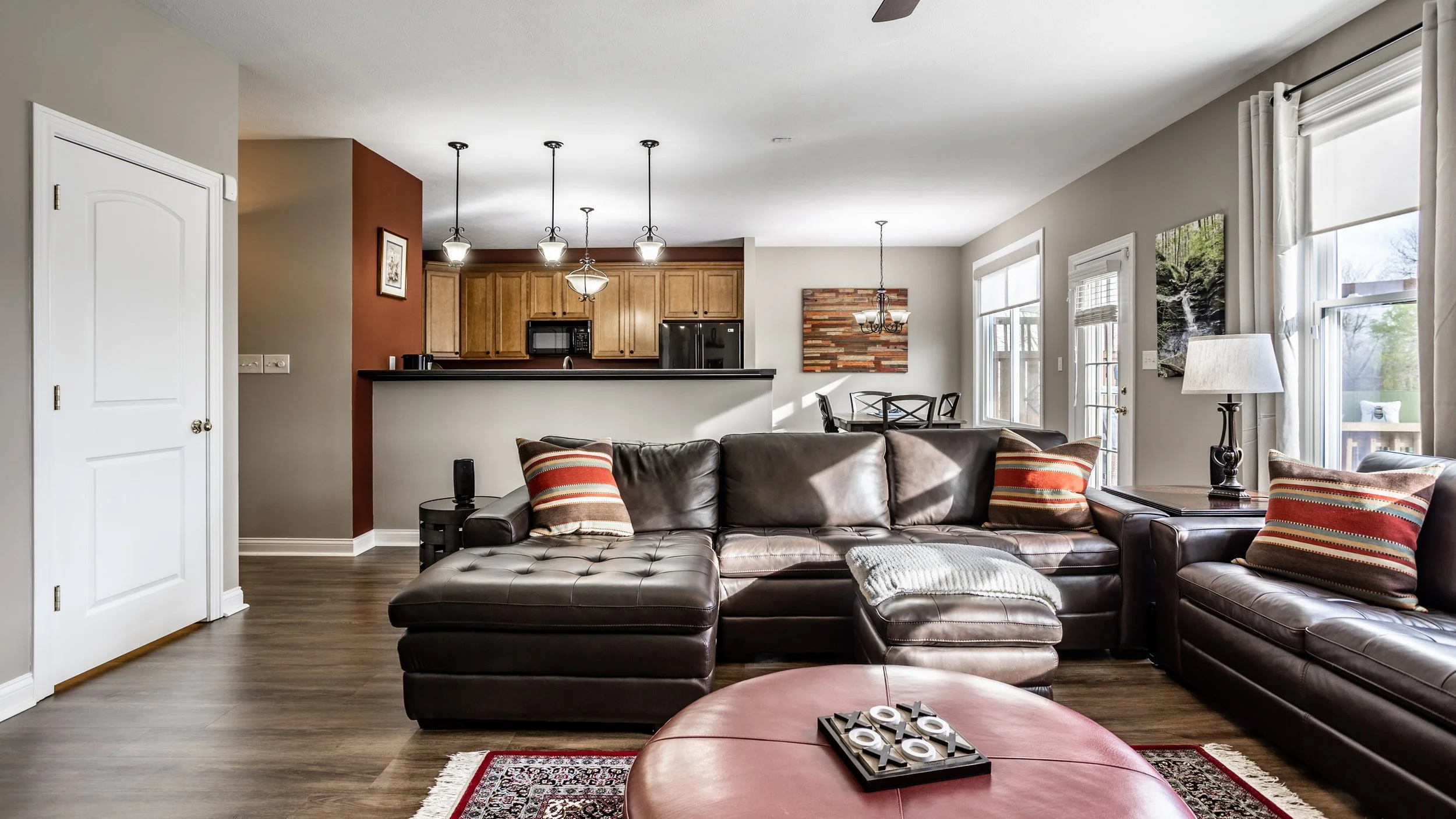 Contemporary living room with brown leather sectional sofa, striped pillows, round leather ottoman, open kitchen with wooden cabinets, pendant lights, dining area, and large windows with blinds.