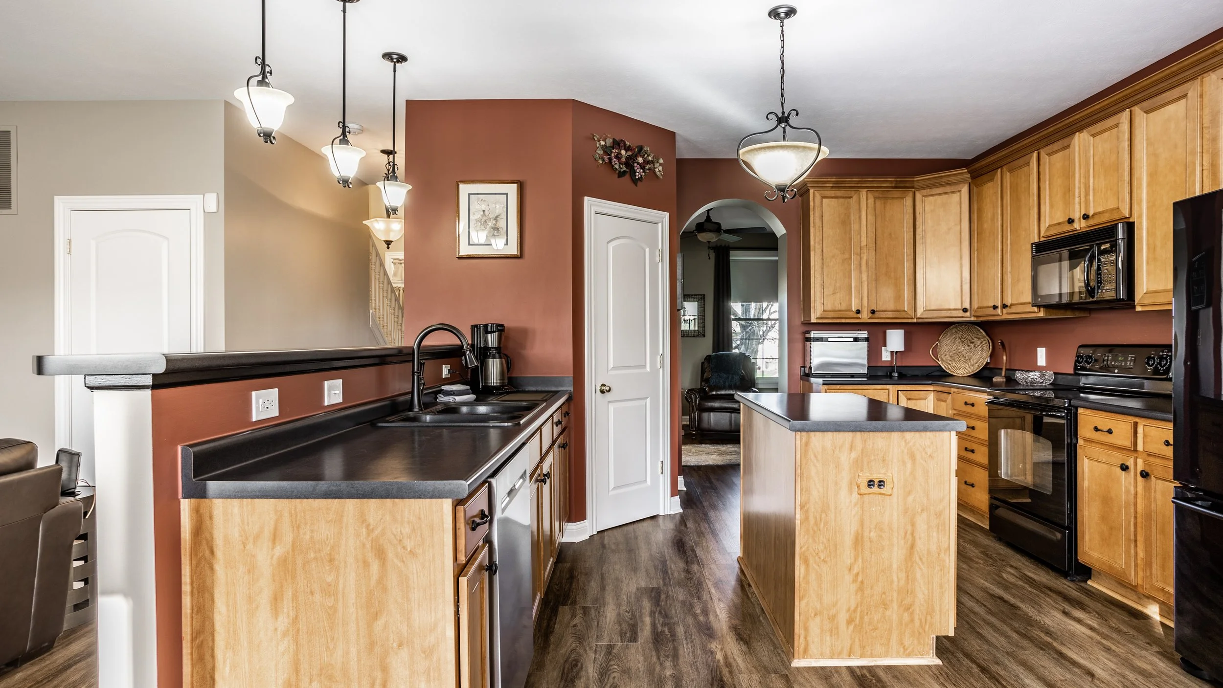 Modern kitchen with wooden cabinets, black countertops, and stainless steel appliances. Pendant lights hang from the ceiling over a central island, while a coffee maker is visible on the counter.
