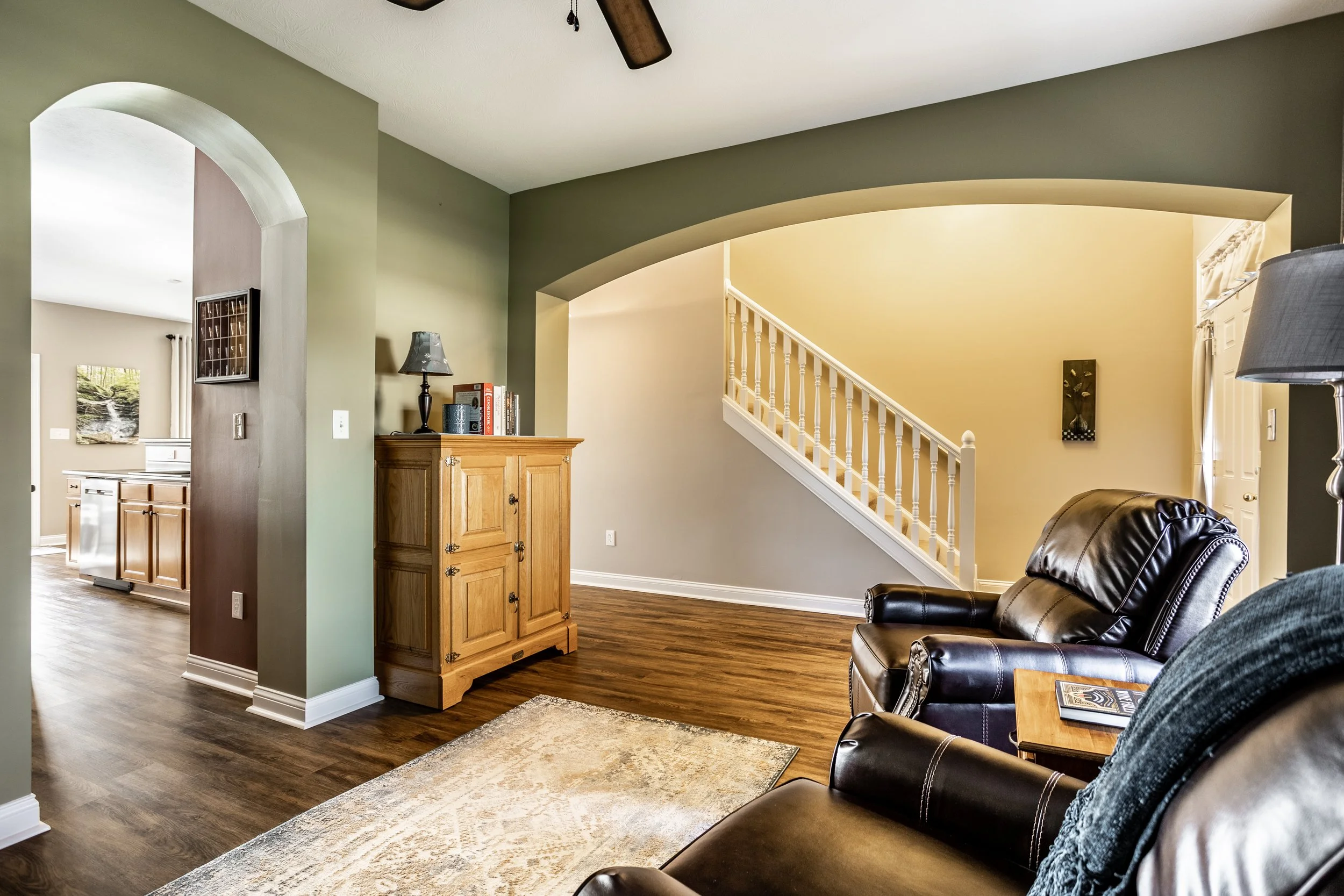Living room with dark leather armchairs, wooden cabinet, archway leading to kitchen, staircase with white railing, and hardwood flooring.