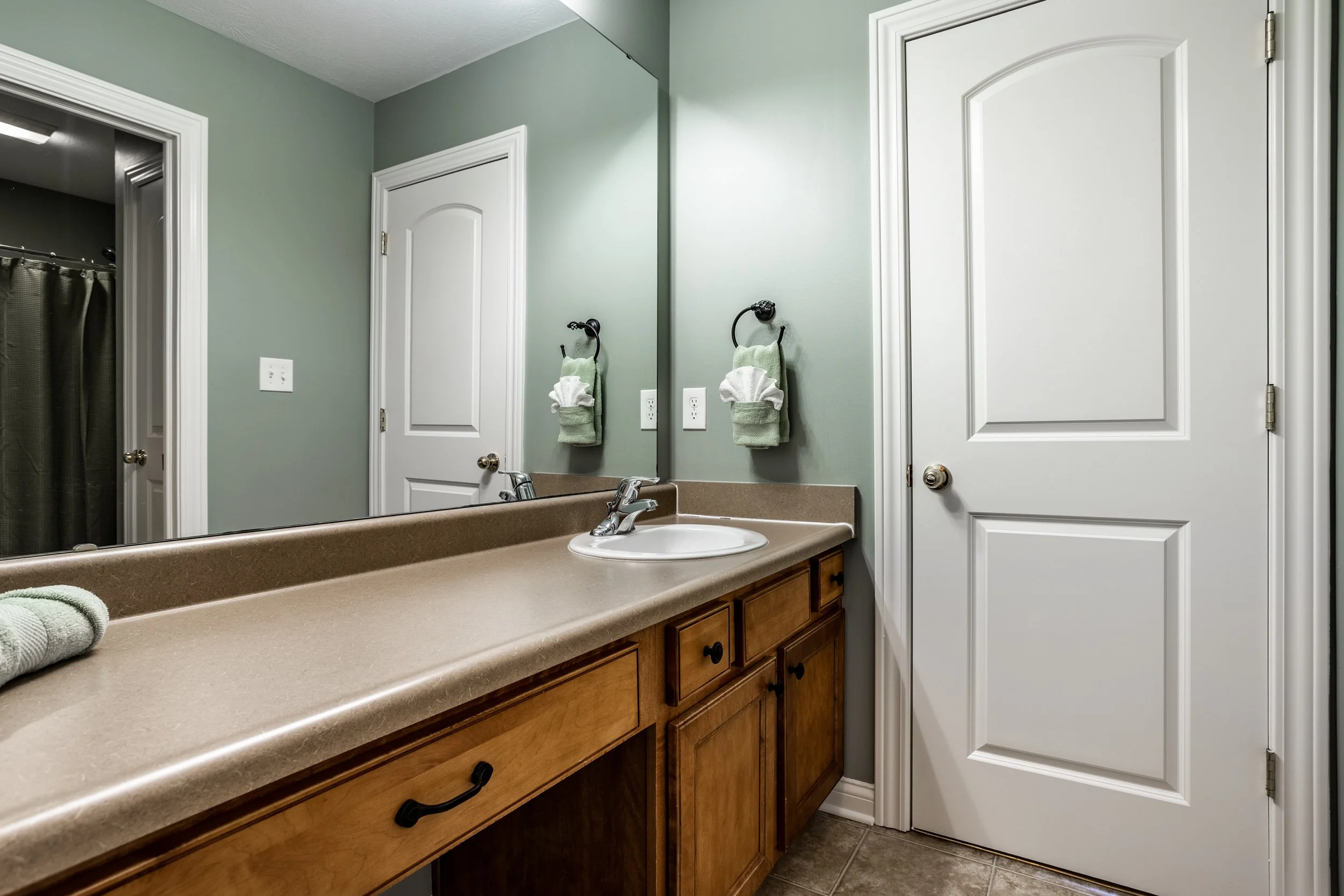 Modern bathroom with green walls, wooden vanity, and white sink. It includes a large mirror, two white doors, and a folded green towel with white accents. Silver faucet and light fixtures are also visible.