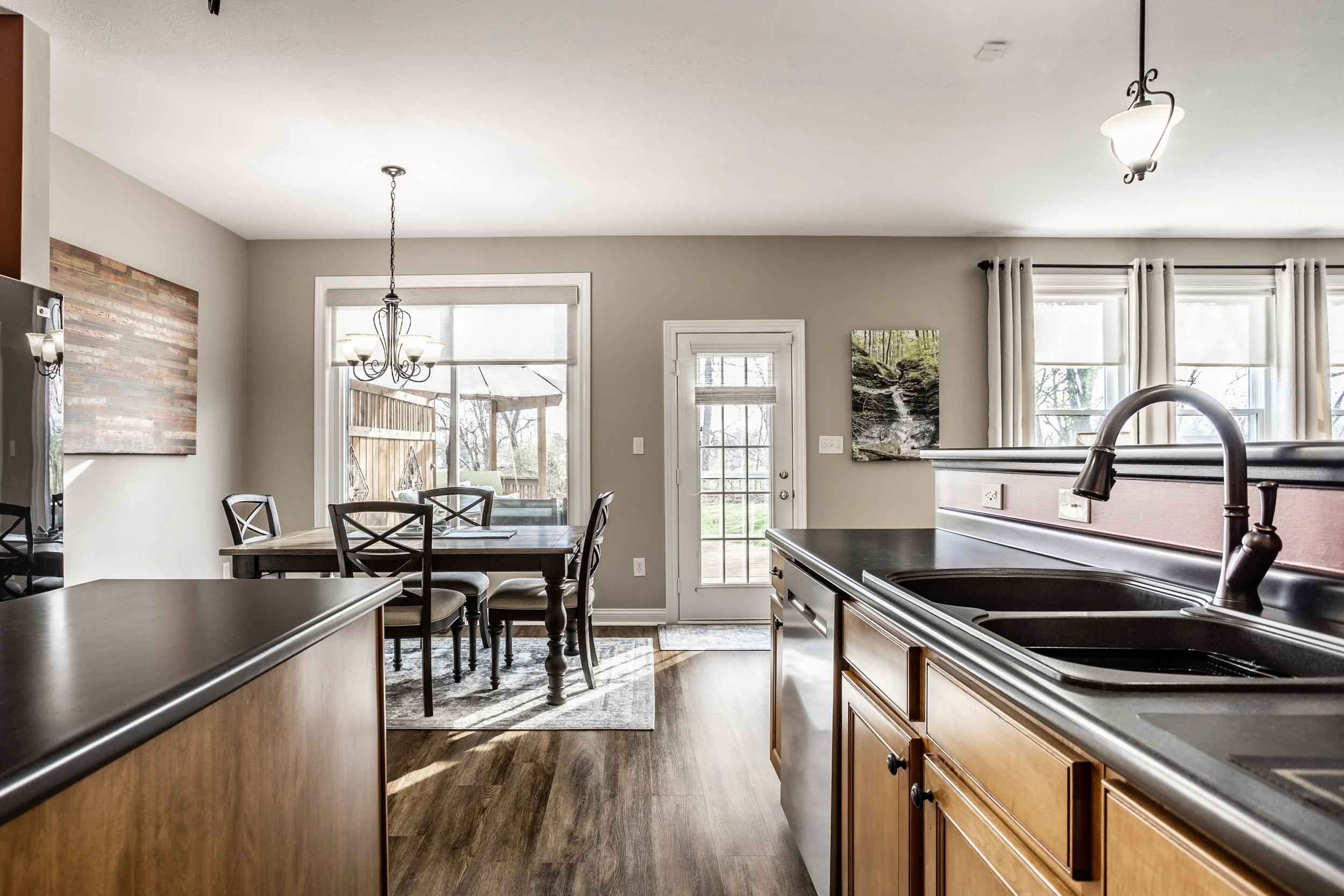 Modern kitchen and dining room with wood cabinets, black countertops, and a dark dining table. A large window and door provide natural light, with a view of the outdoor deck. A chandelier hangs above the dining area.