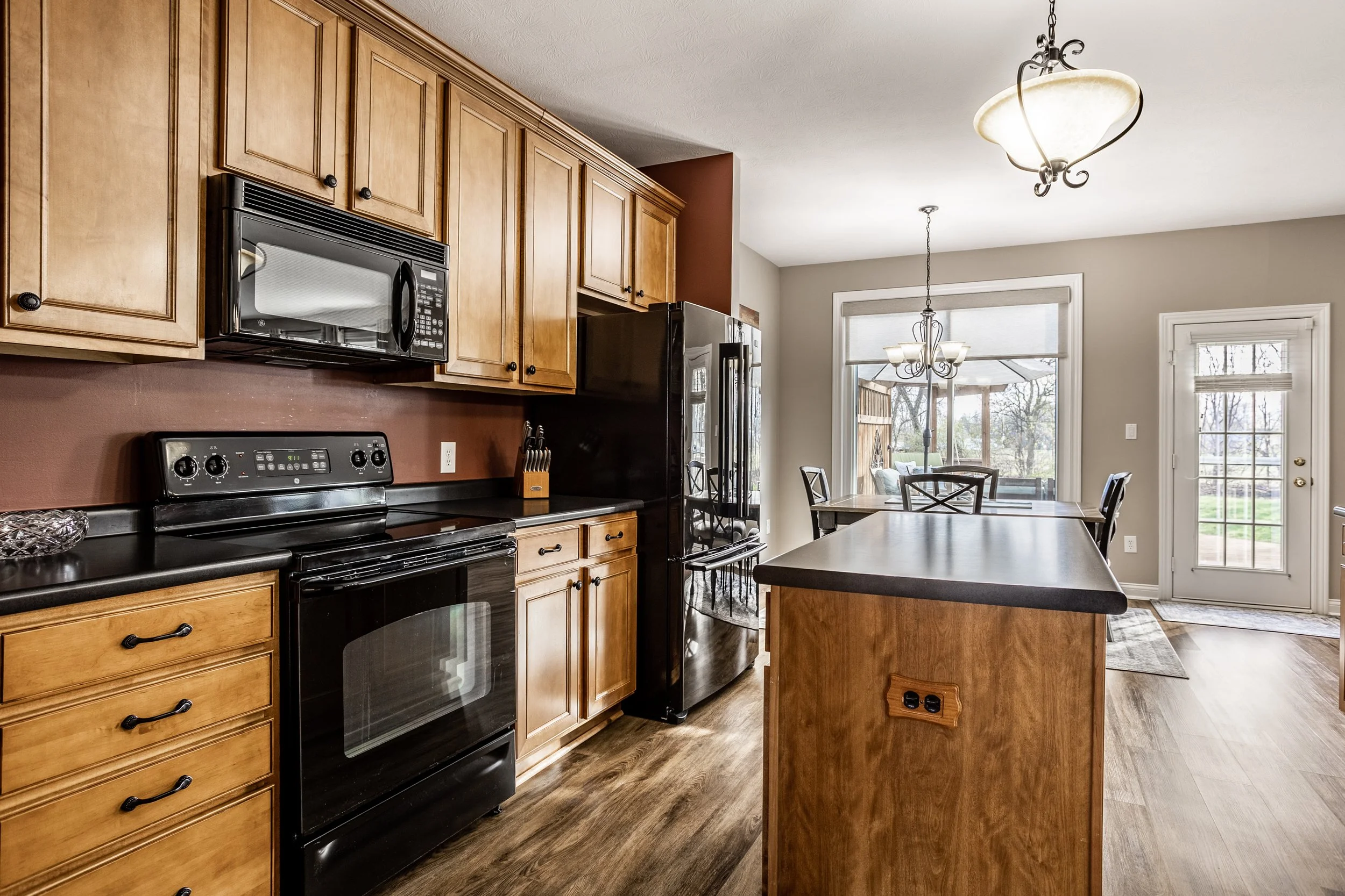Modern kitchen with wooden cabinets, black appliances, and large island. Features a stove, microwave, refrigerator, and light fixtures. Open layout with dining area and outdoor view through glass doors.