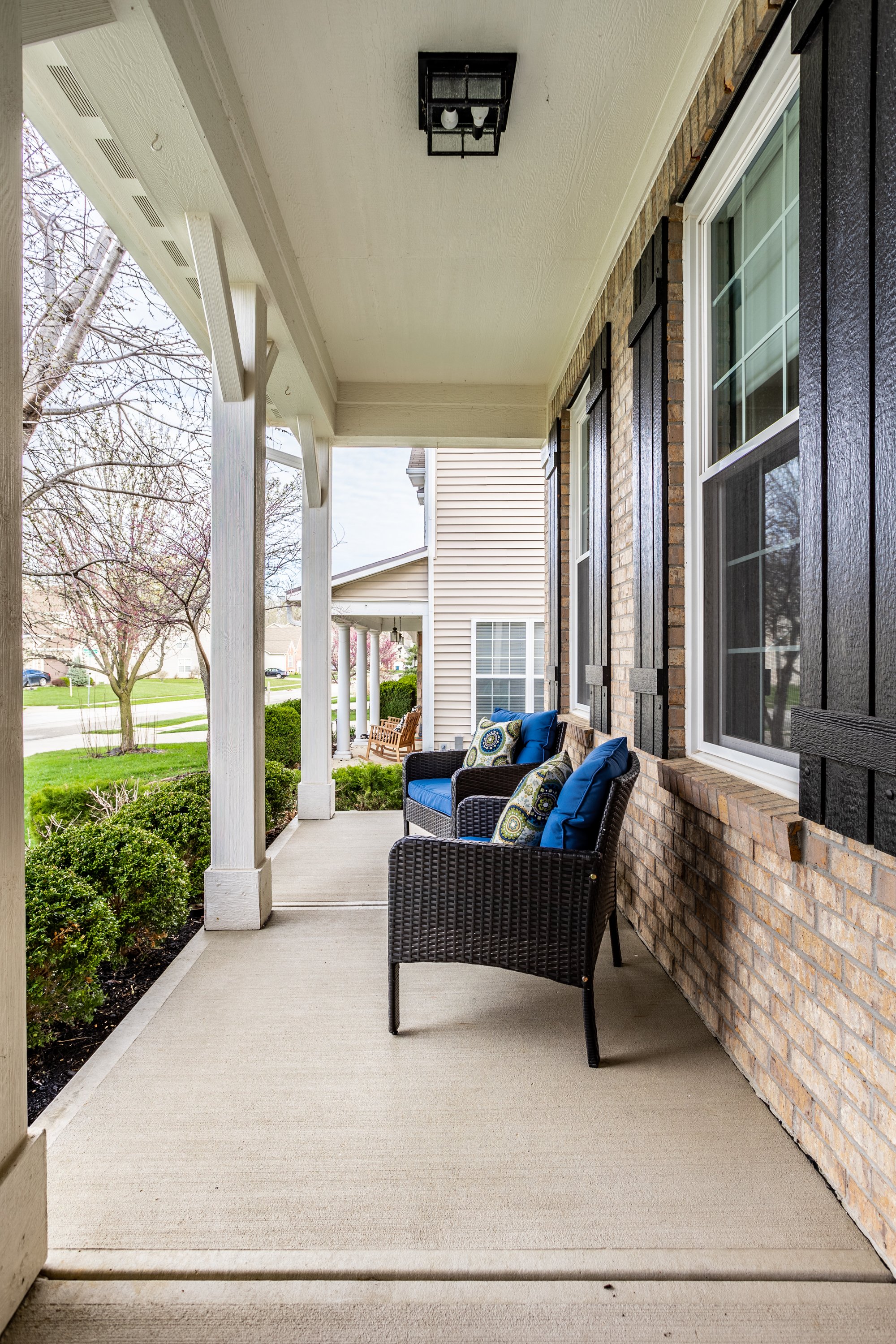 Front porch with black wicker chairs, blue cushions, decorative pillows, brick exterior wall, white columns, and greenery.