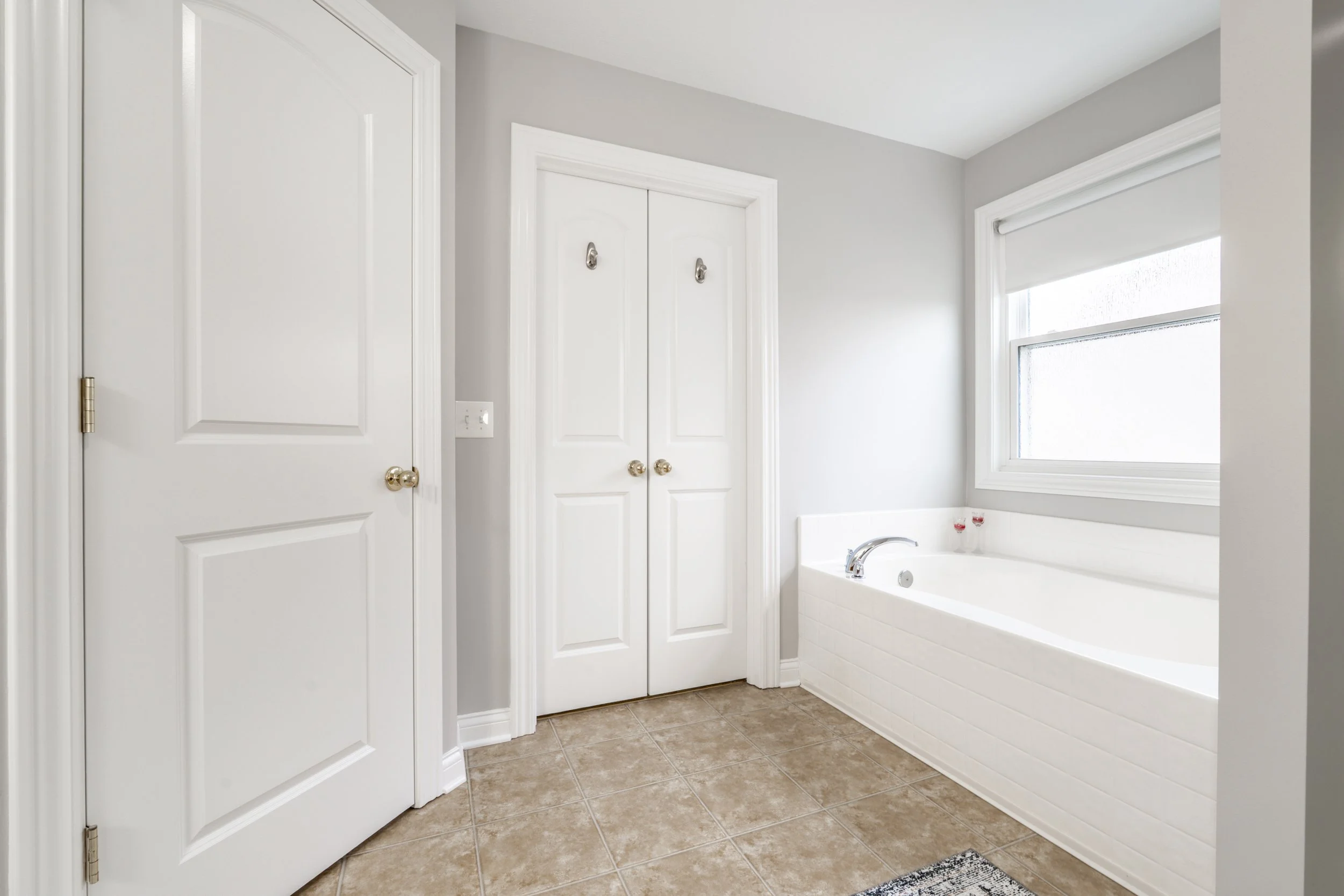Modern bathroom with white bathtub, tiled floor, closed double doors, and a frosted window.