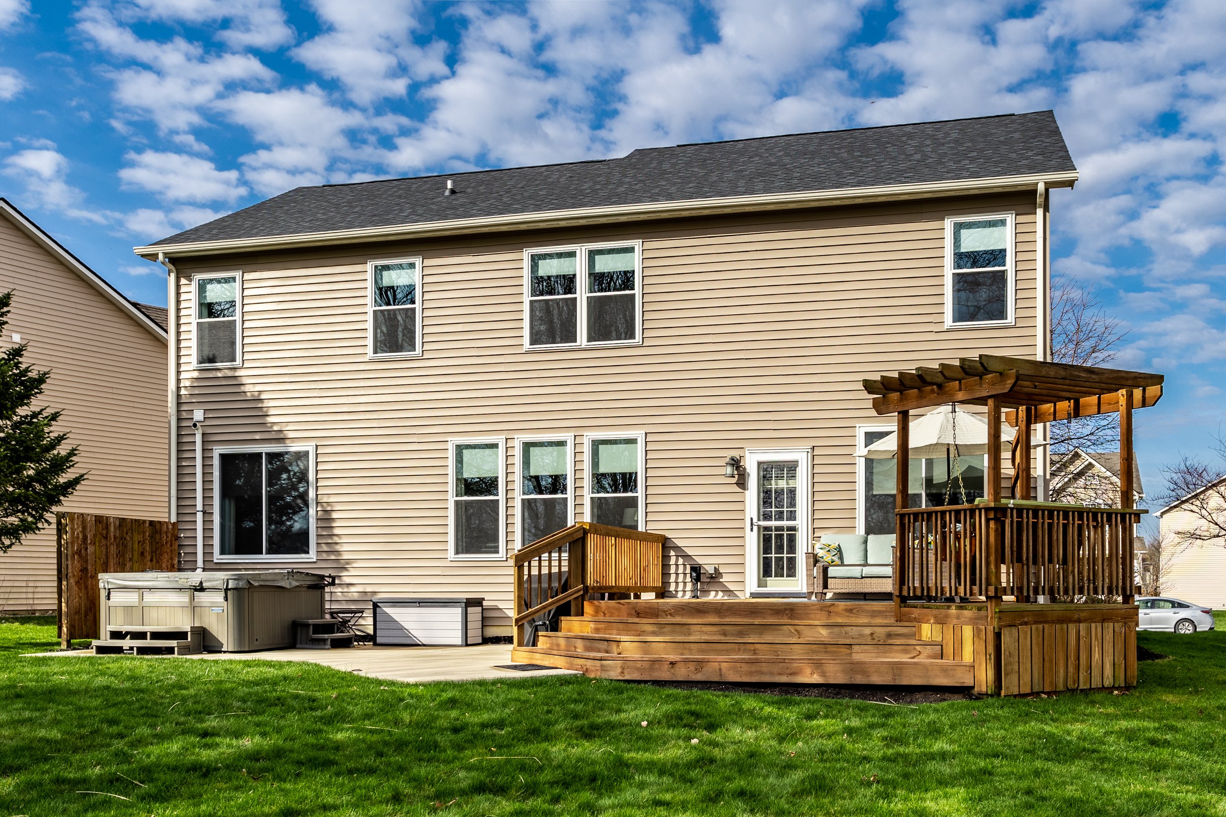 Backyard view of a suburban house with beige siding, featuring a wooden deck and pergola, a hot tub, green lawn, and partial view of neighboring houses under a blue sky with clouds.
