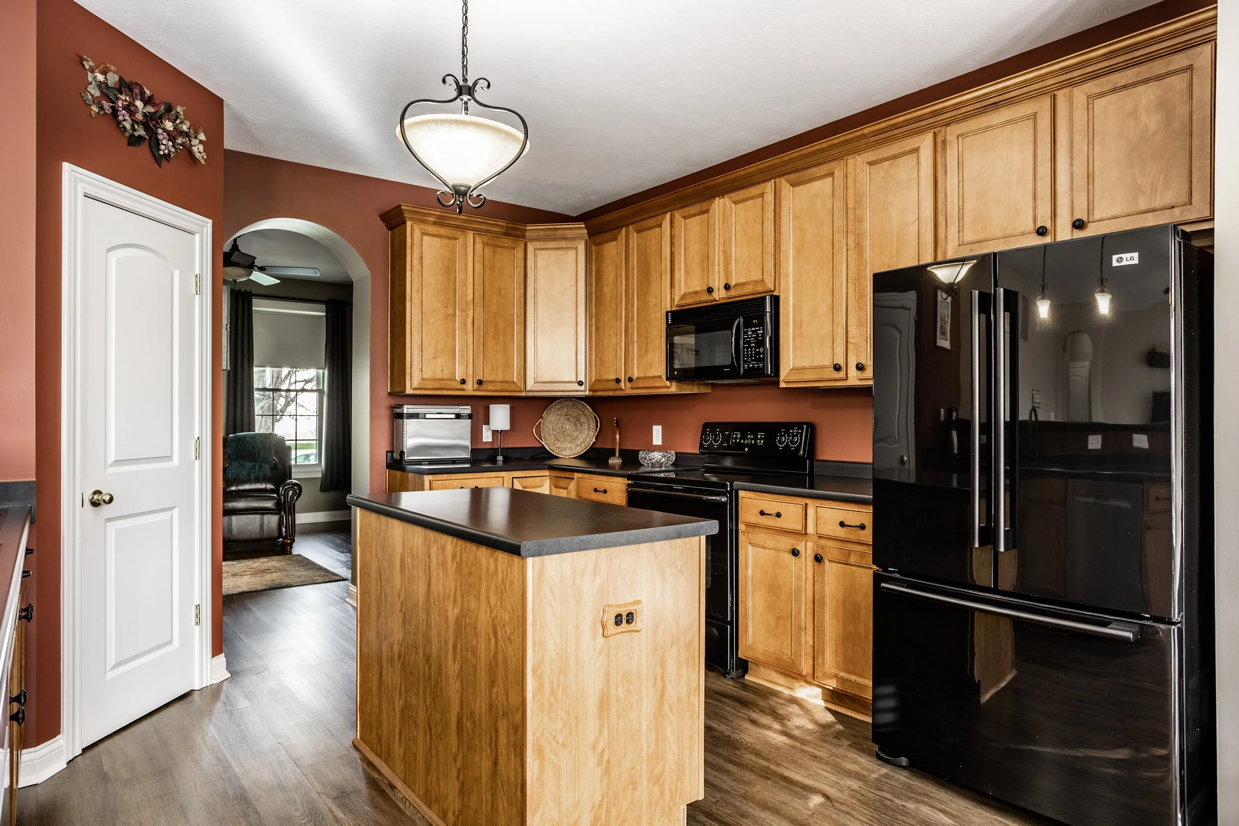 Modern kitchen with wooden cabinets, black appliances, island, and dark countertops, featuring a pendant light, hardwood floor, and archway leading to another room.