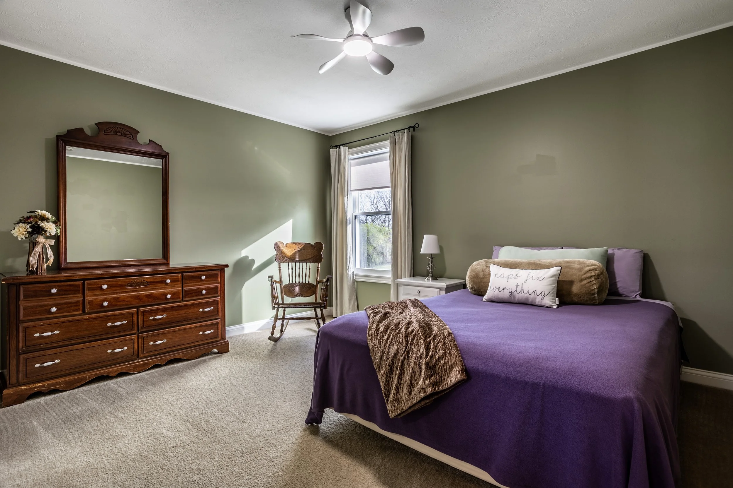 Bedroom with green walls, purple bedspread, wooden dresser with mirror, rocking chair, bedside table, and a ceiling fan.