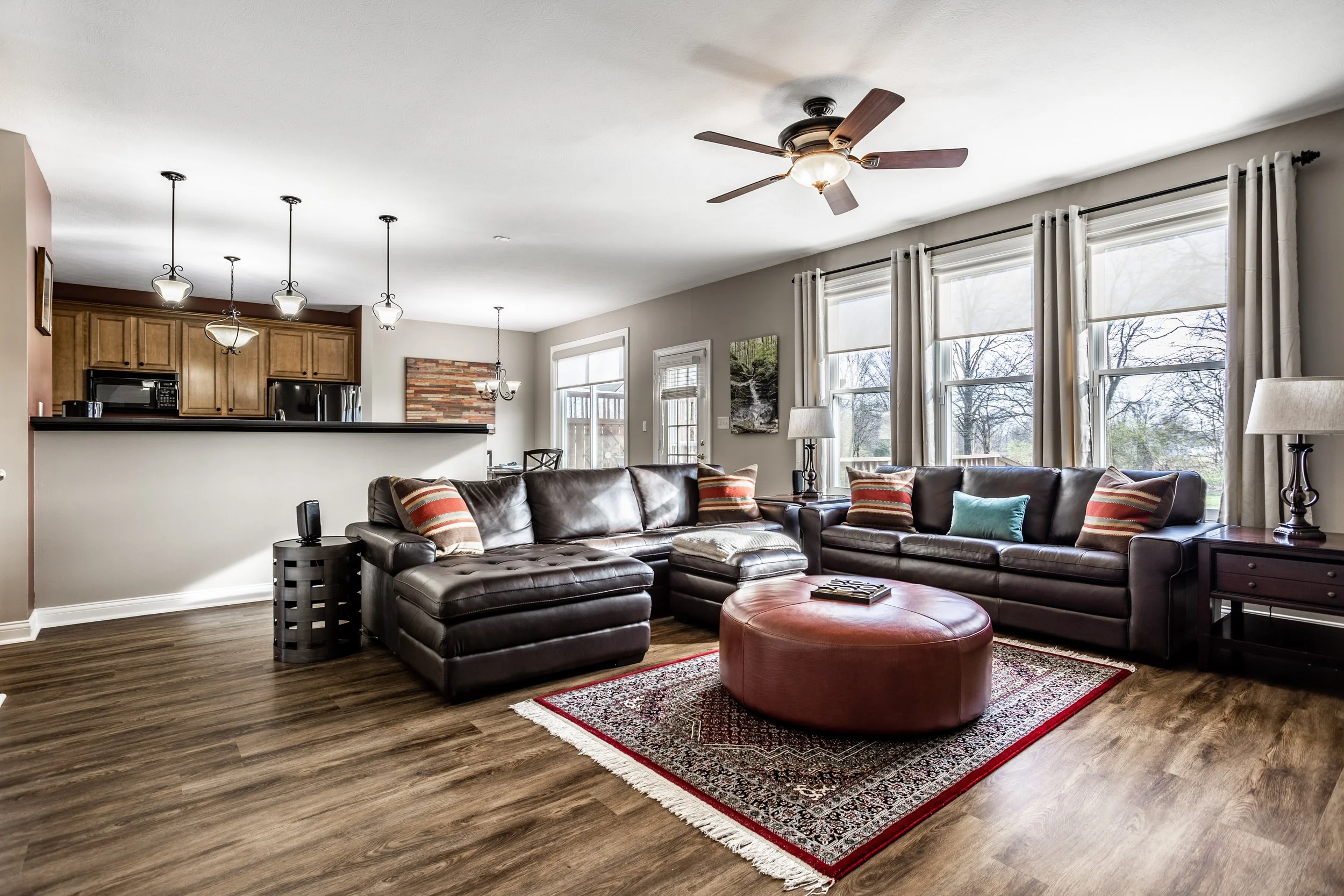 Contemporary living room with brown leather sofas, colorful cushions, a large red ottoman, wood flooring, and open kitchen with pendant lighting.