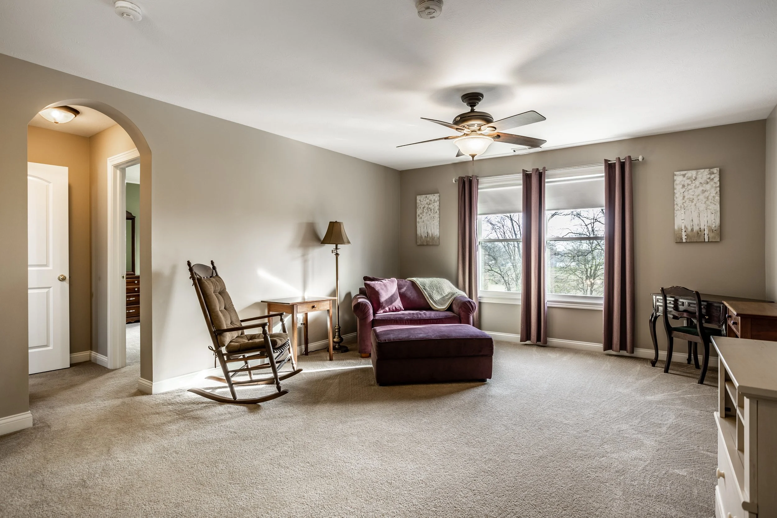 Spacious living room with a purple armchair and ottoman, rocking chair, wooden desk, and large windows with curtains. Beige carpet and walls, ceiling fan, and natural light.