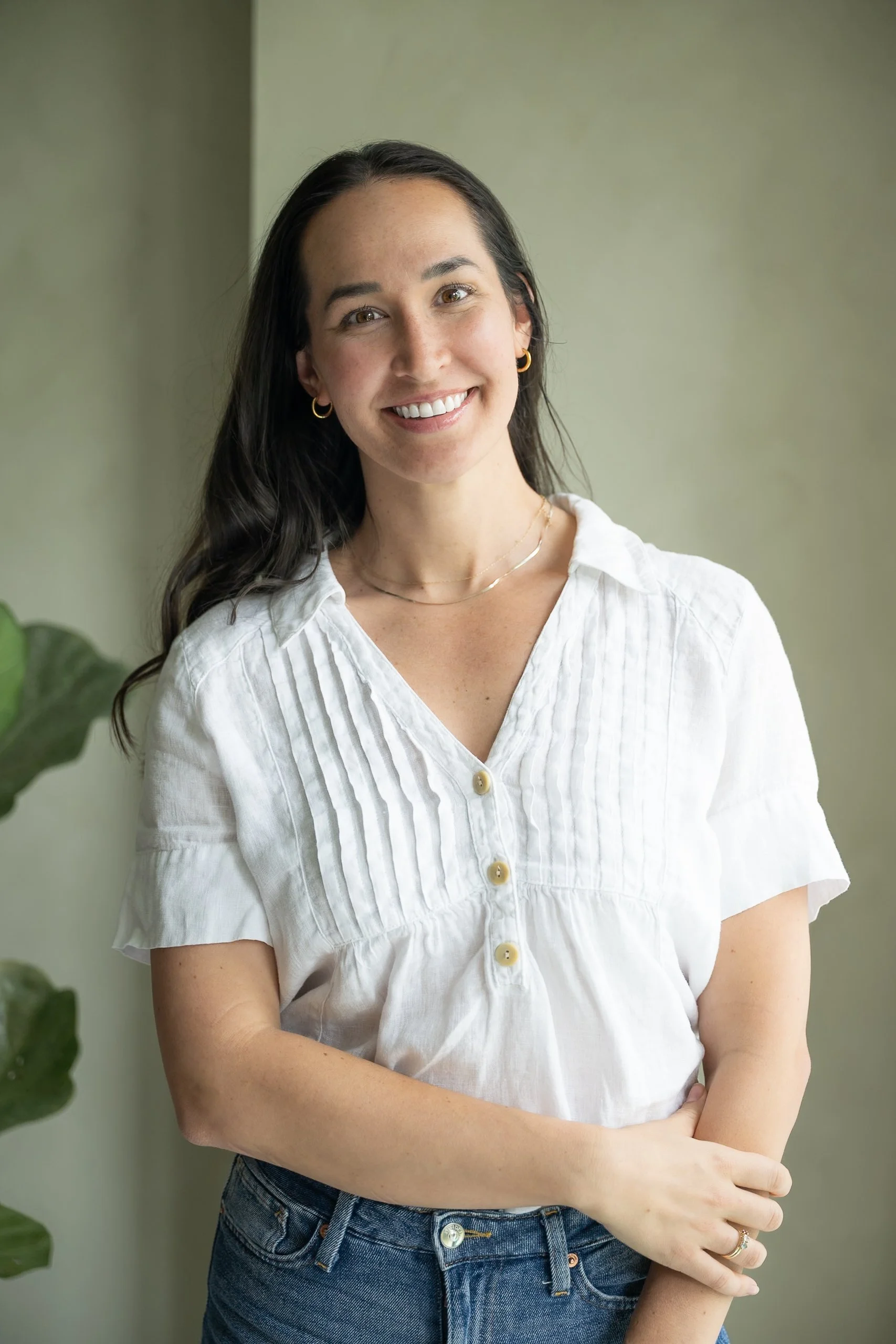 A woman with long dark hair, smiling, wearing a white button-up blouse and blue jeans, standing indoors near a light-colored wall, with a green plant partially visible to her left.