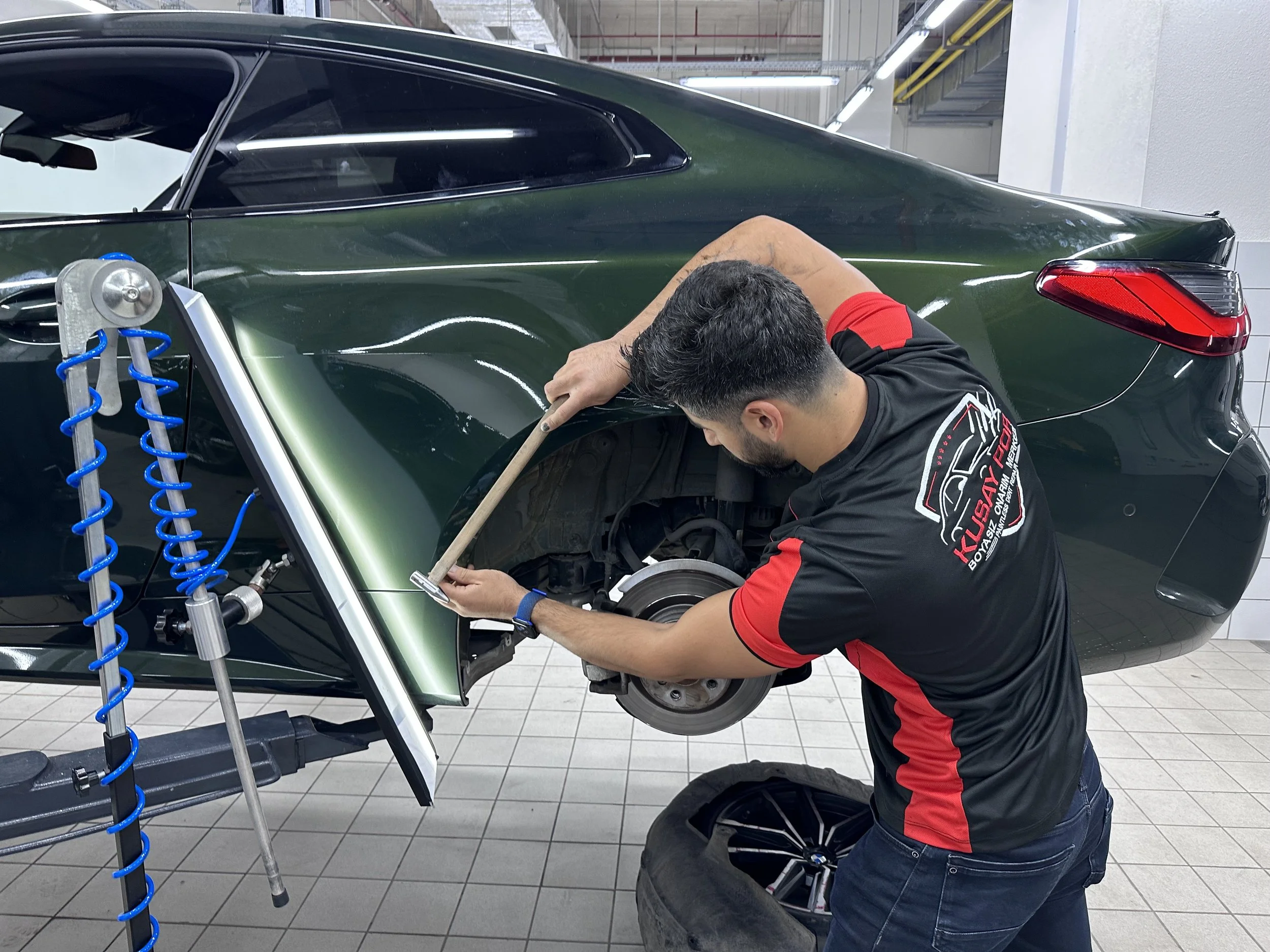 A technician repairing the rear side of a green car, using a tool to fix dents. The car is elevated with the wheel removed. An alignment tool with a blue coil is visible beside the car.