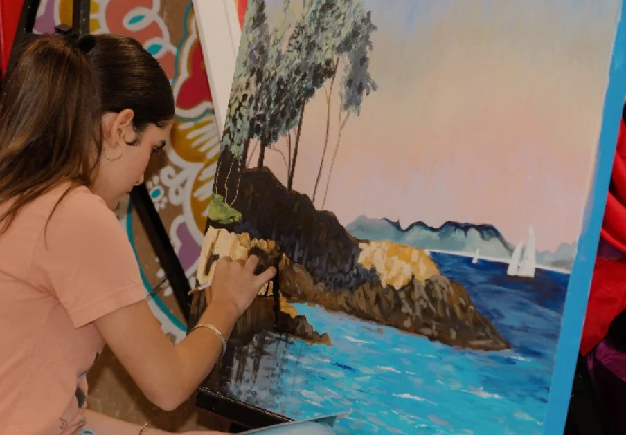 A young girl painting a seascape with rocks, trees, and sailboats.