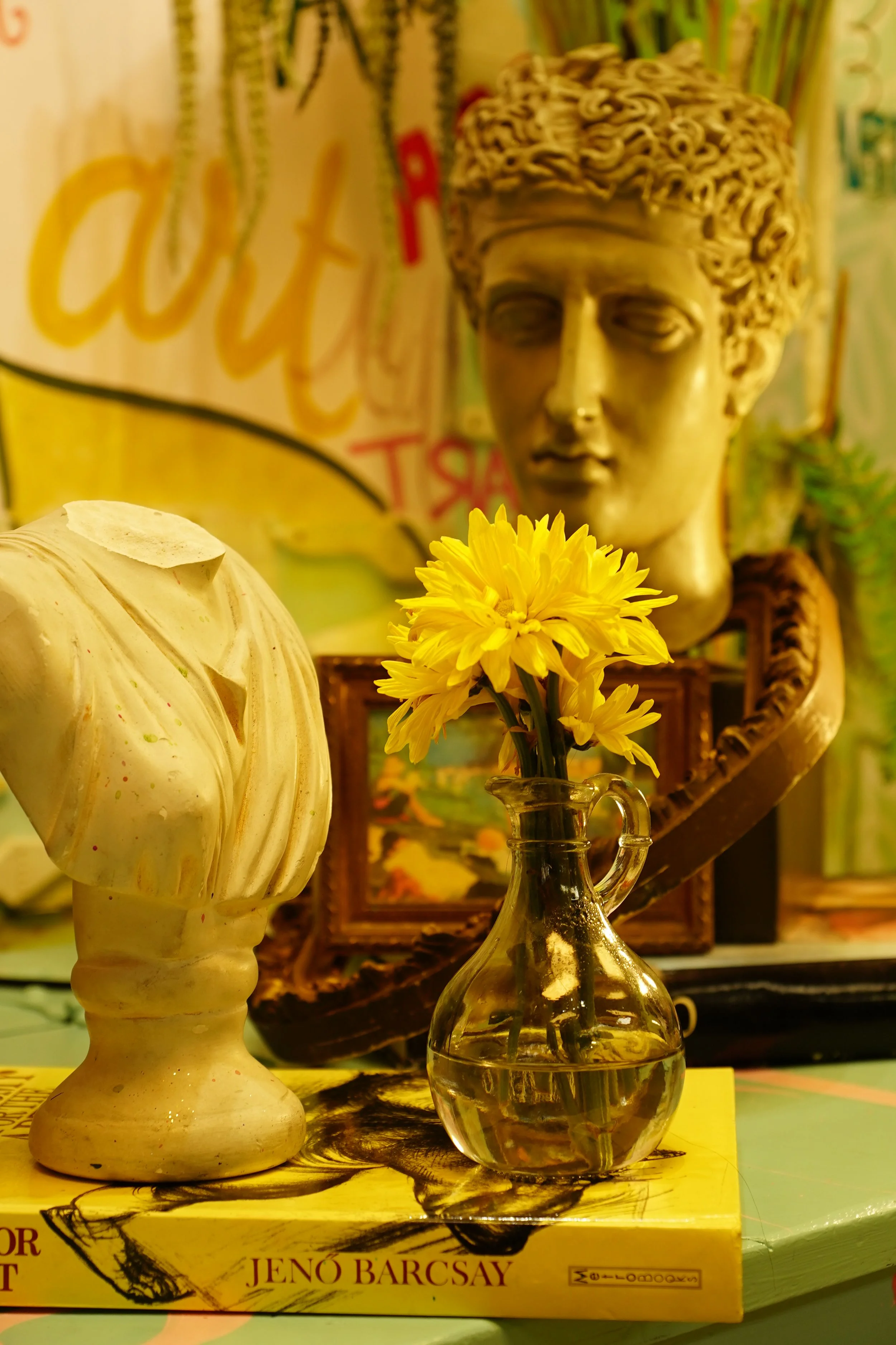 A decorative arrangement featuring a neoclassical bust, a yellow flower bouquet in a glass vase, and an art book by Jenő Barcsay. The items are set against a colorful backdrop with artistic elements.