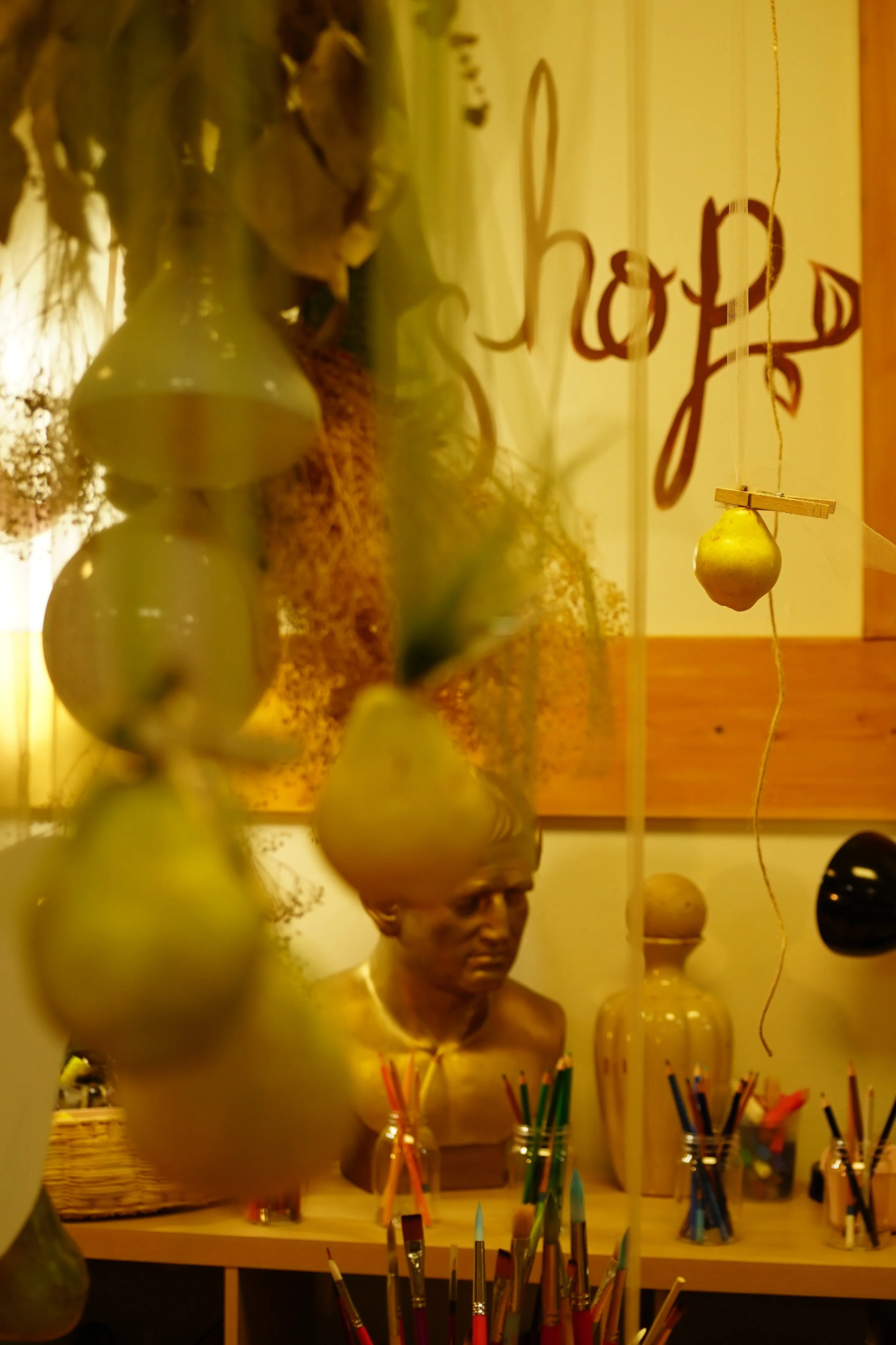 Art studio interior with a bust sculpture, hanging pears, jars of colored pencils, and the word 'shop' on the wall.