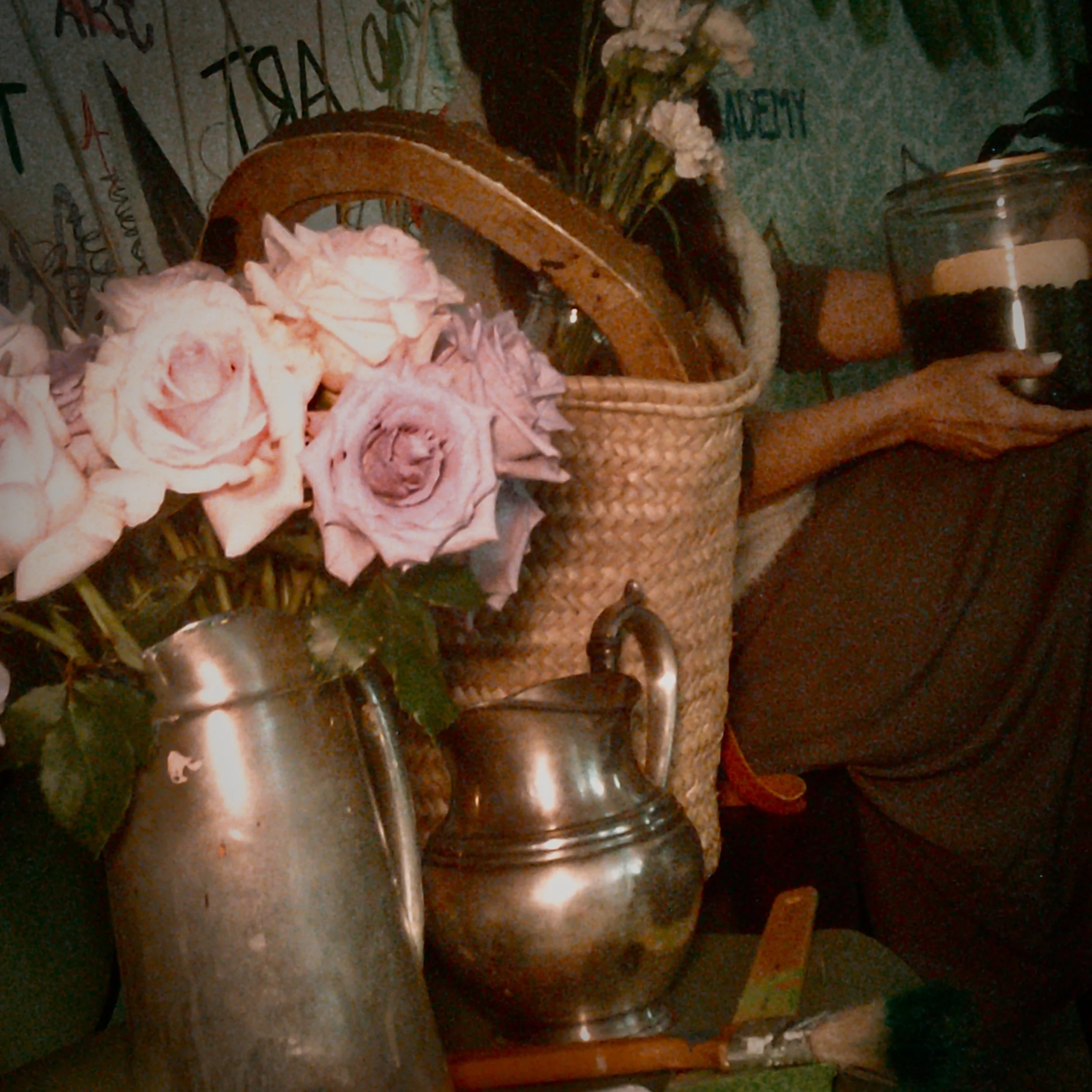 A bouquet of roses in a metal pitcher, next to a woven basket and a silver jug, with a person holding a jar in the background.
