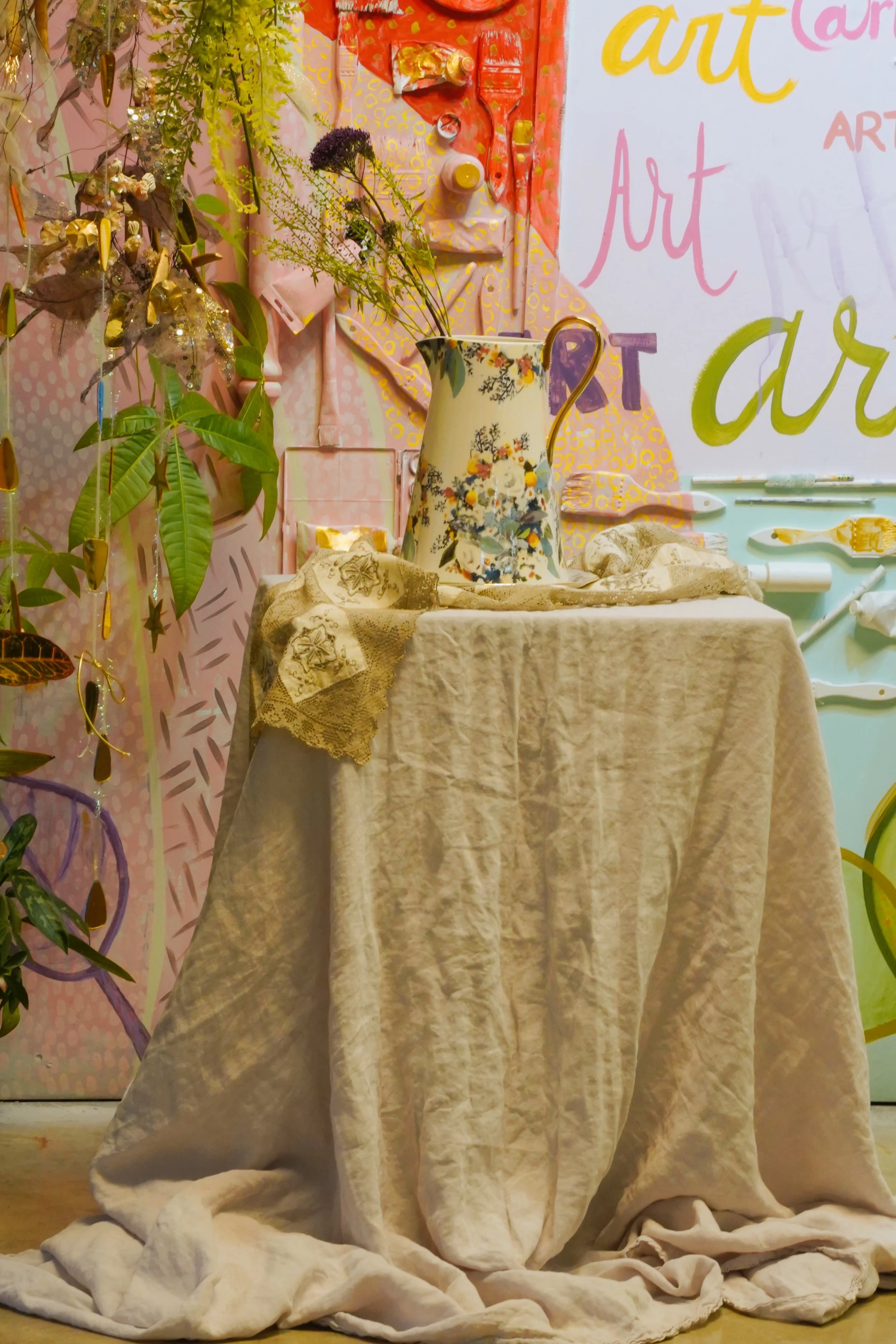 Decorative floral pitcher on table with beige cloth, surrounded by artistic background and greenery.