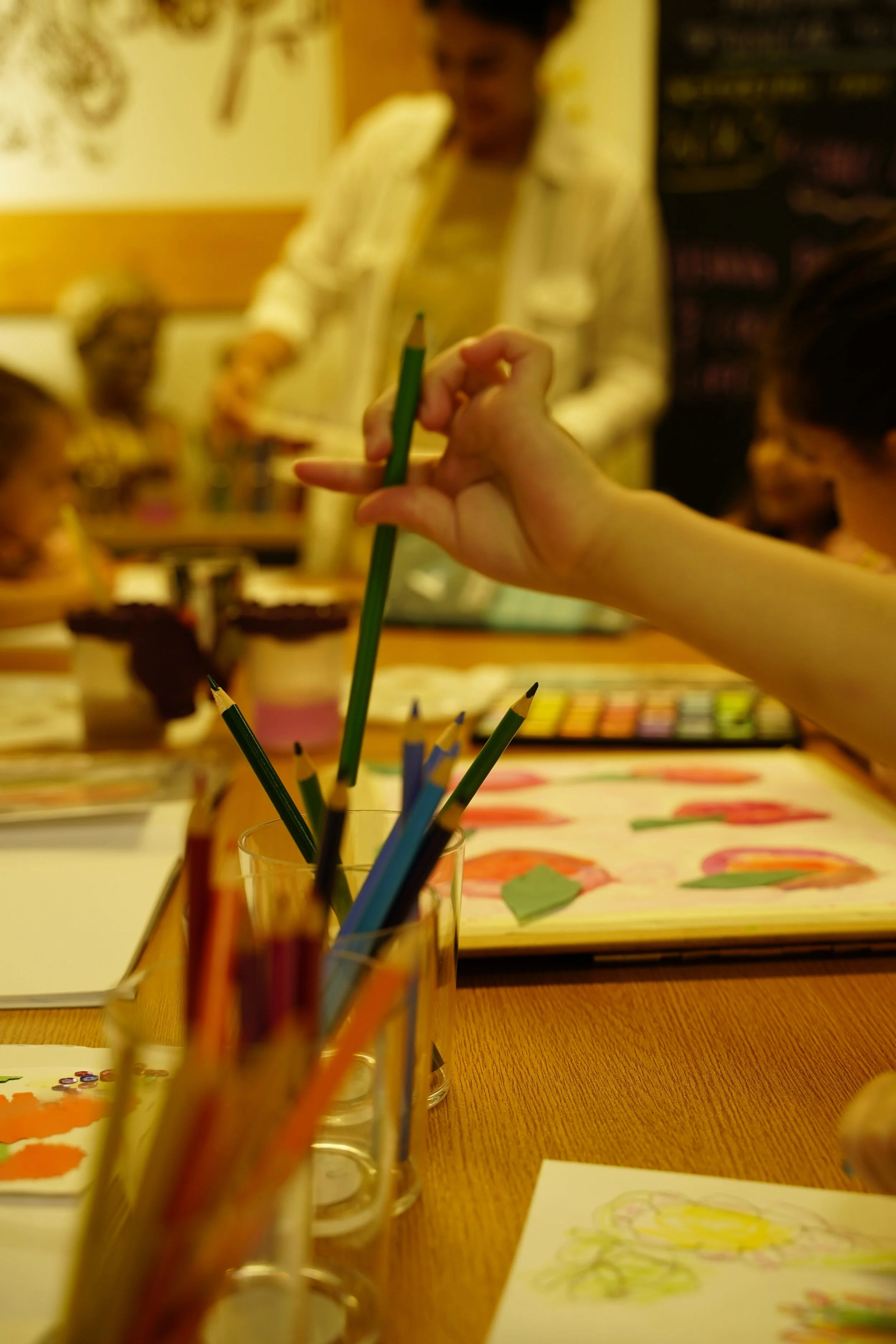 A person holding a green colored pencil in an art classroom, with other colored pencils in a glass. Children are drawing colorful pictures in the background, with art supplies on the wooden table.