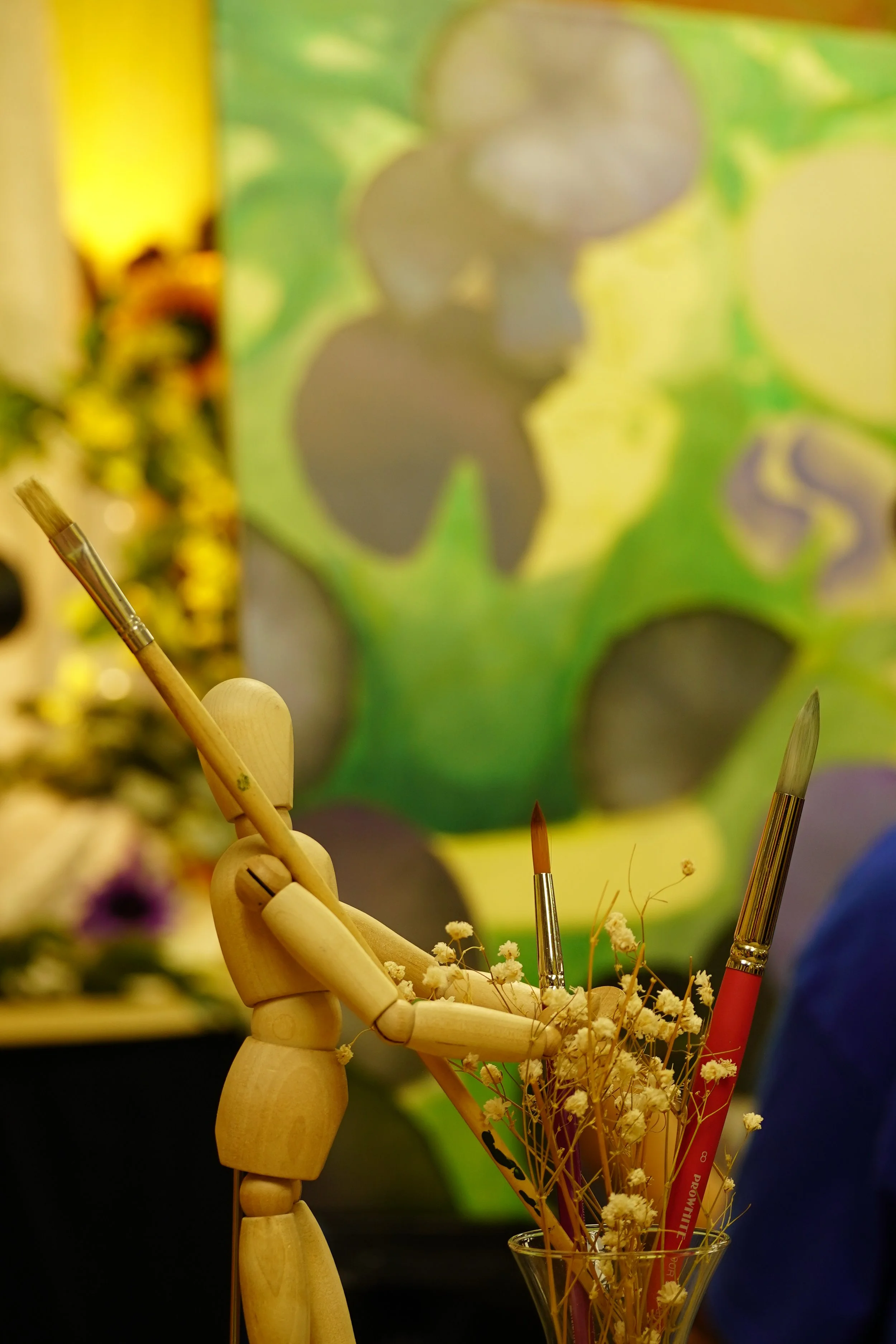 Wooden mannequin posing with paintbrushes, flowers, and abstract green and purple painting in background.