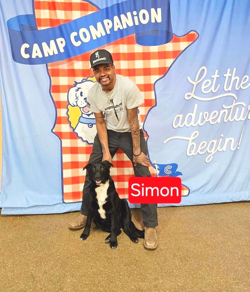 A man and a black and white dog sitting together at an event with a backdrop that says 'Camp Companion' and 'Let the adventure begin!'. The man is smiling and the dog's name is Simon.