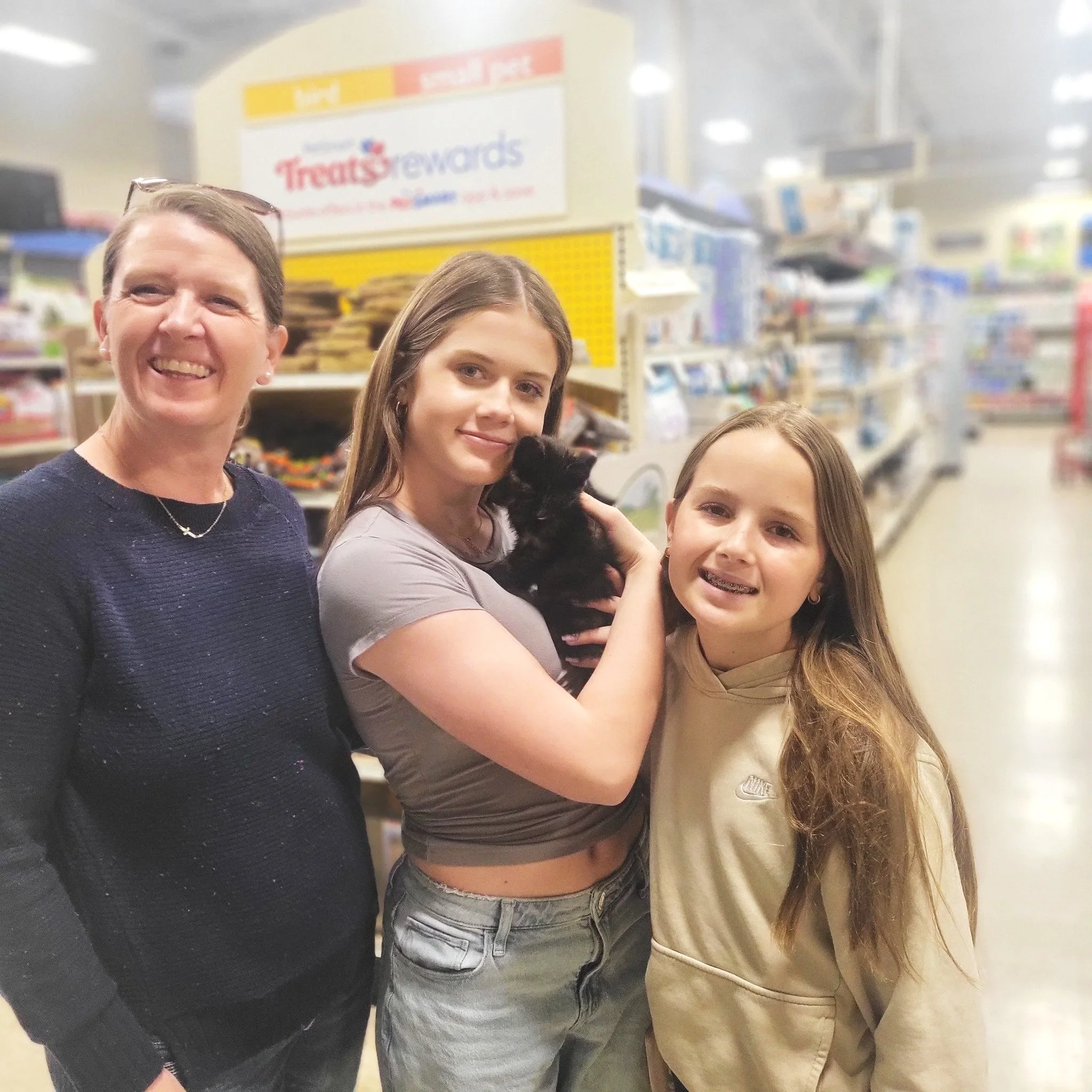 Three smiling gals in a pet store with one holding a black kitten.