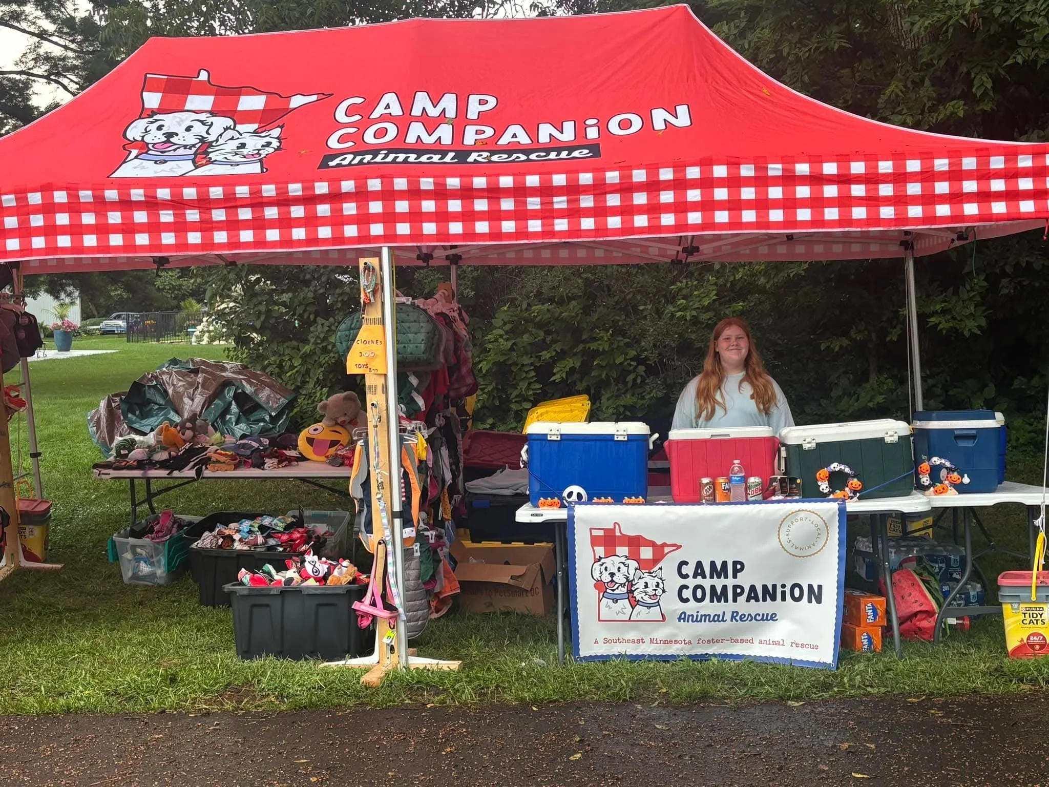 A red tent with the words "Camp Companion Animal Rescue" and under the tent there are coolers, toys, and items for sale, with a young girl smiling behind the table. The tent is set up outdoors on a grassy area.