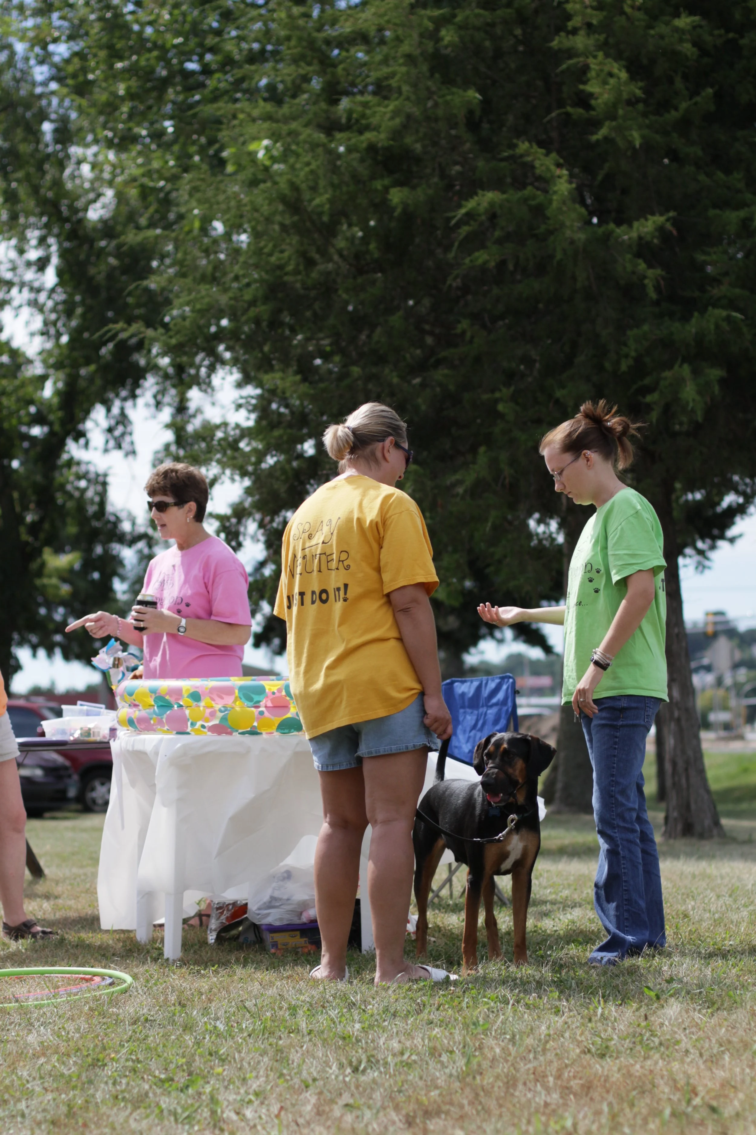 People gathered outdoors at a park for a dog-related event, with a woman holding a Rottweiler, a table with food, and large trees in the background.