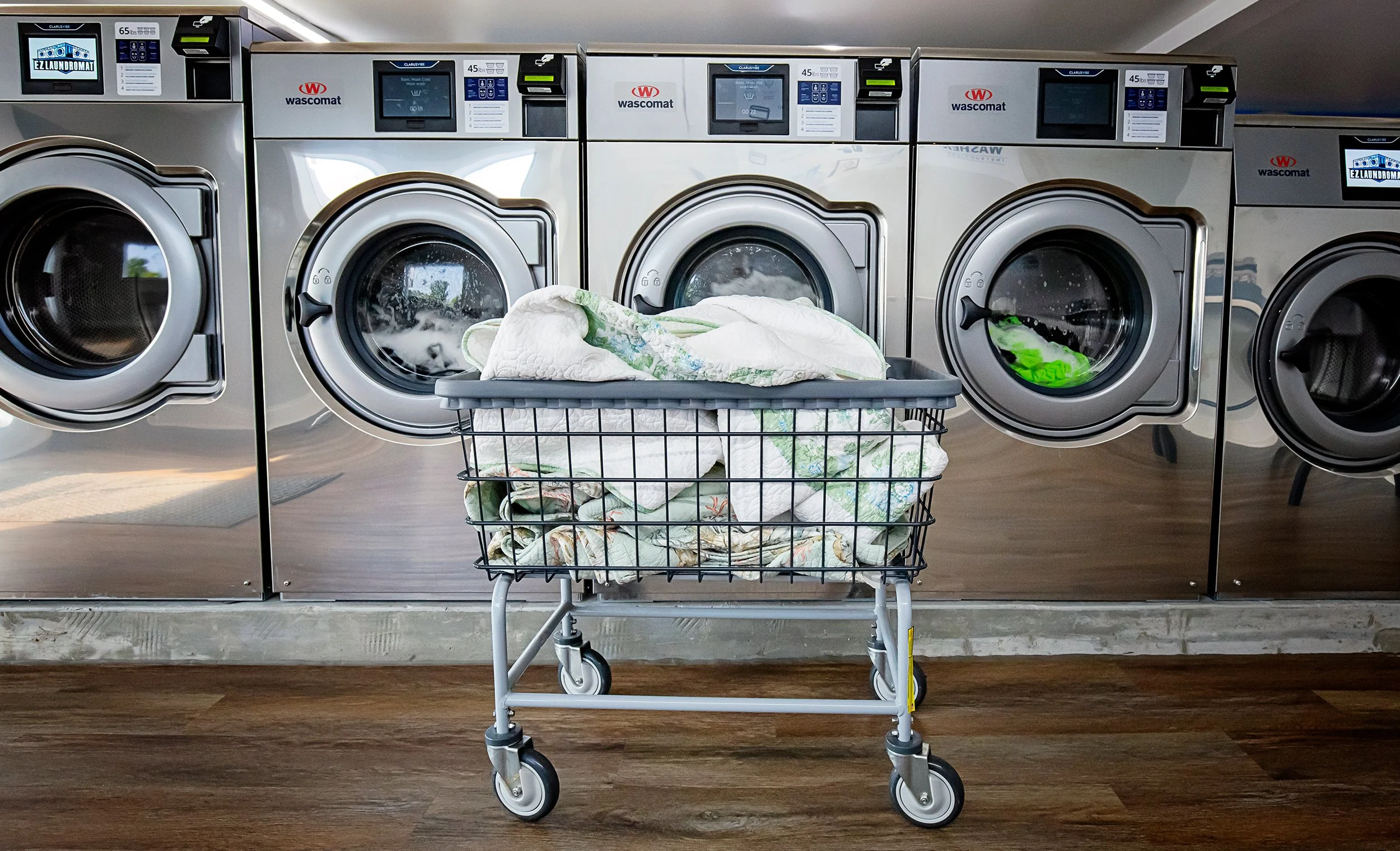 A laundry room with four washing machines and a shopping cart filled with laundry in front of the machines.