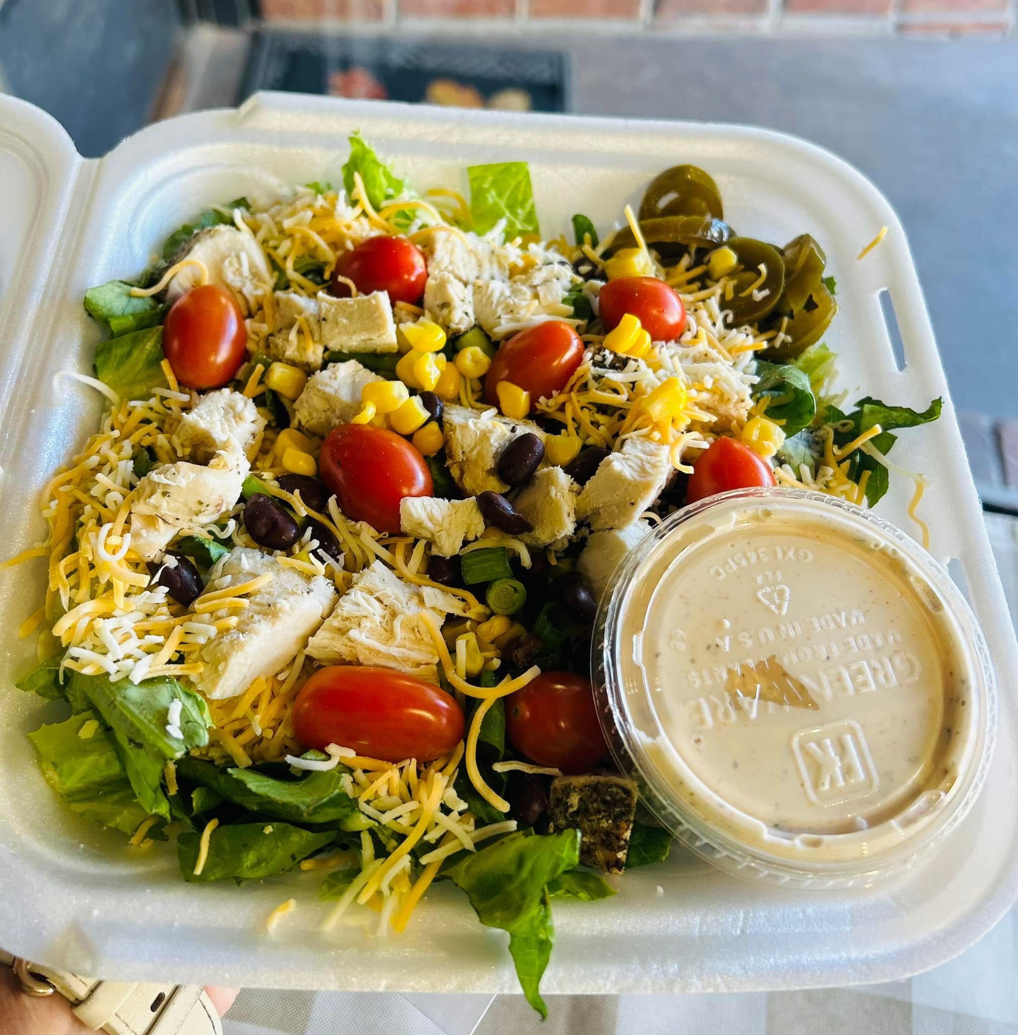 A chicken salad with cherry tomatoes, shredded cheese, corn, black beans, jalapeños, and lettuce in a takeout container with a side of creamy dressing.