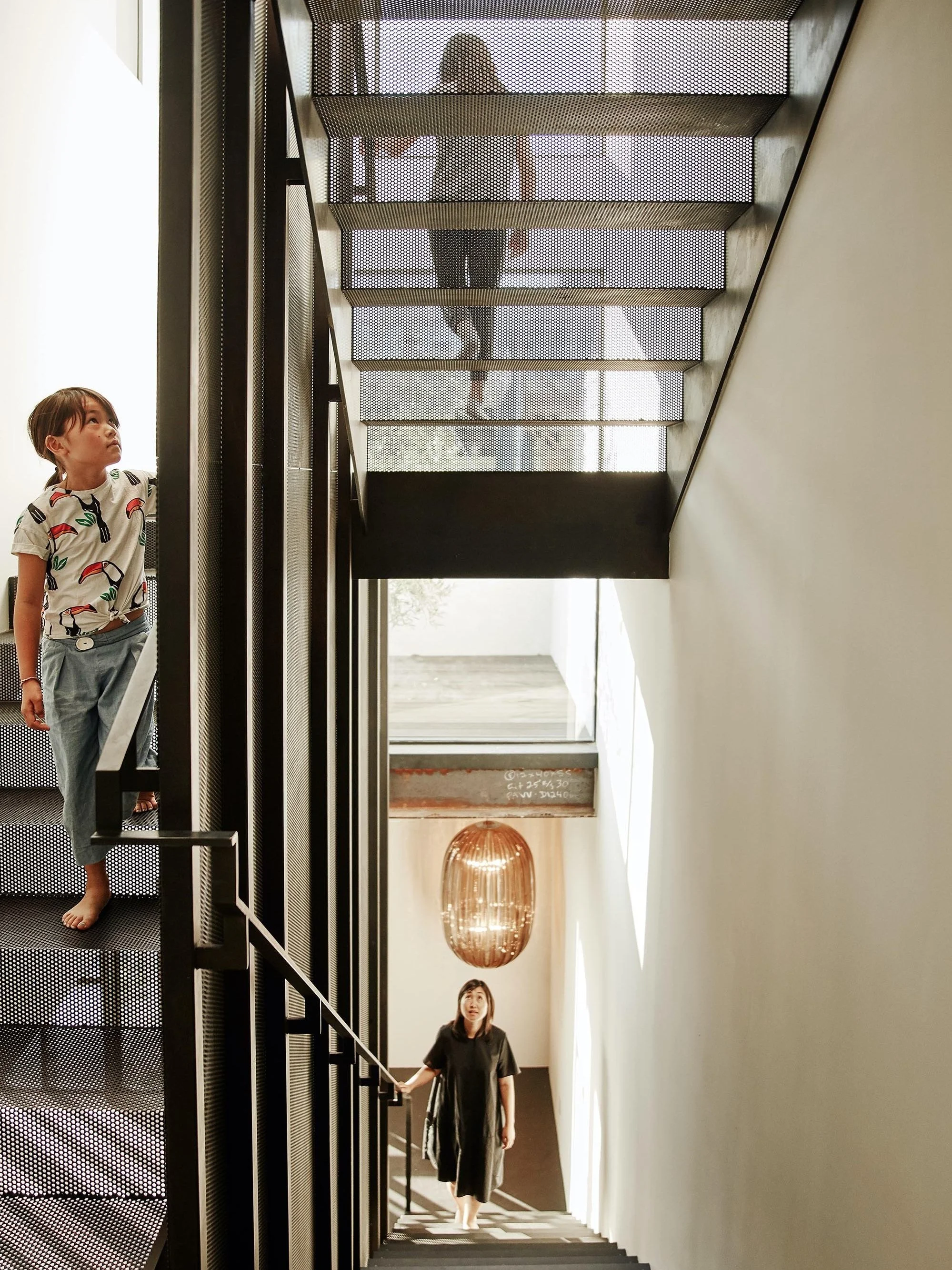 Two people walking on a modern metal staircase, with one person ascending and another descending. A large hanging lamp is visible in the background.