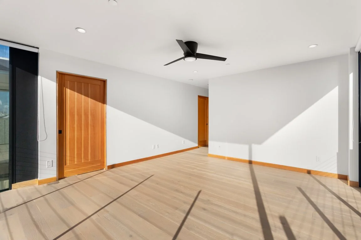 Empty room with white walls, hardwood flooring, wooden doors, black ceiling fan, and sunlight casting shadows.