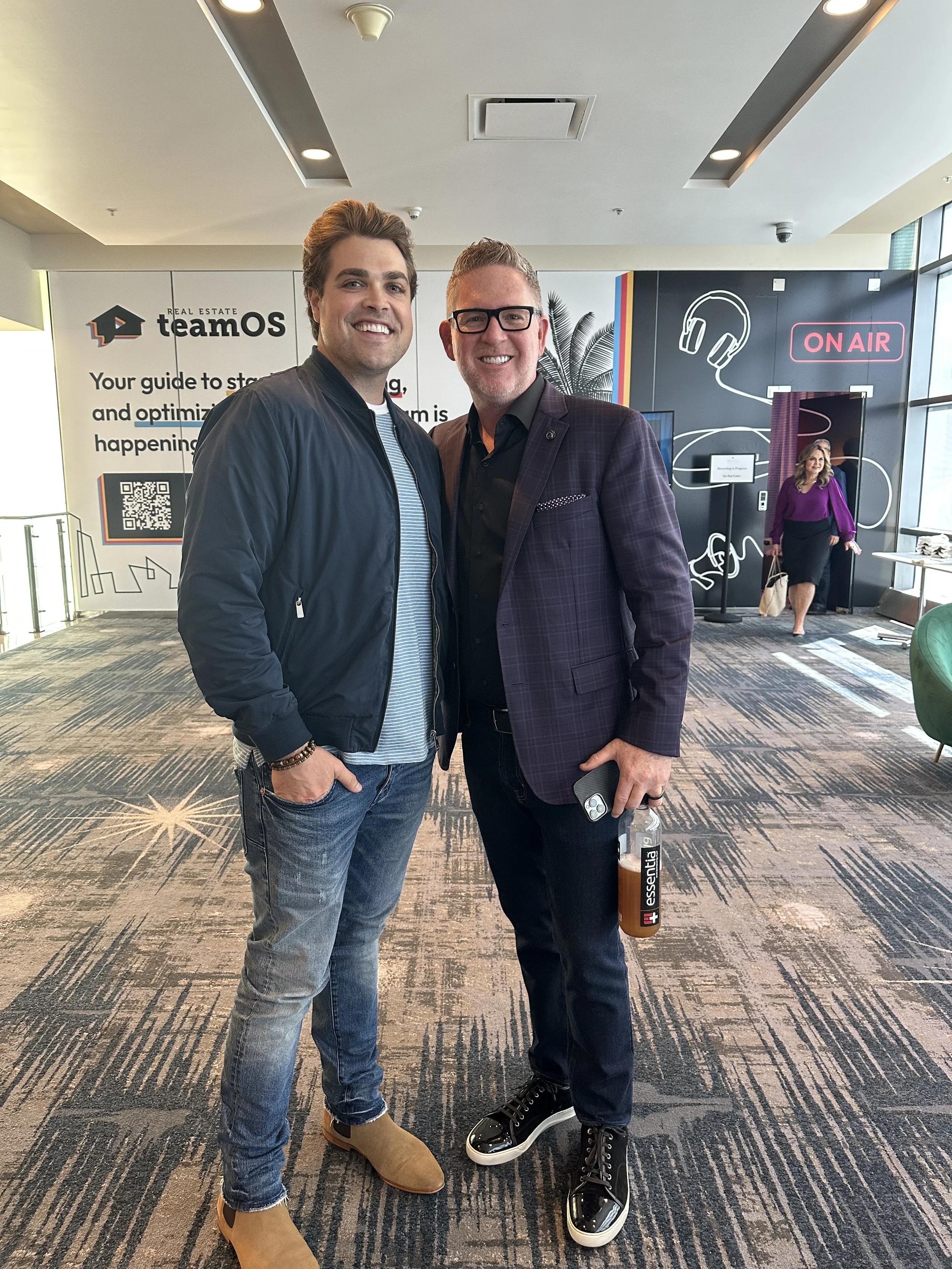 Two men standing together inside a modern office or conference space, smiling at the camera. One is wearing a black jacket and jeans, the other a dark plaid blazer and black pants, holding a drink and a phone. In the background, there is a woman walk