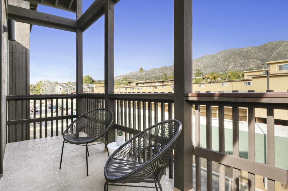 Balcony with two black chairs, railing, overlooking neighboring buildings and distant hills under a clear blue sky.