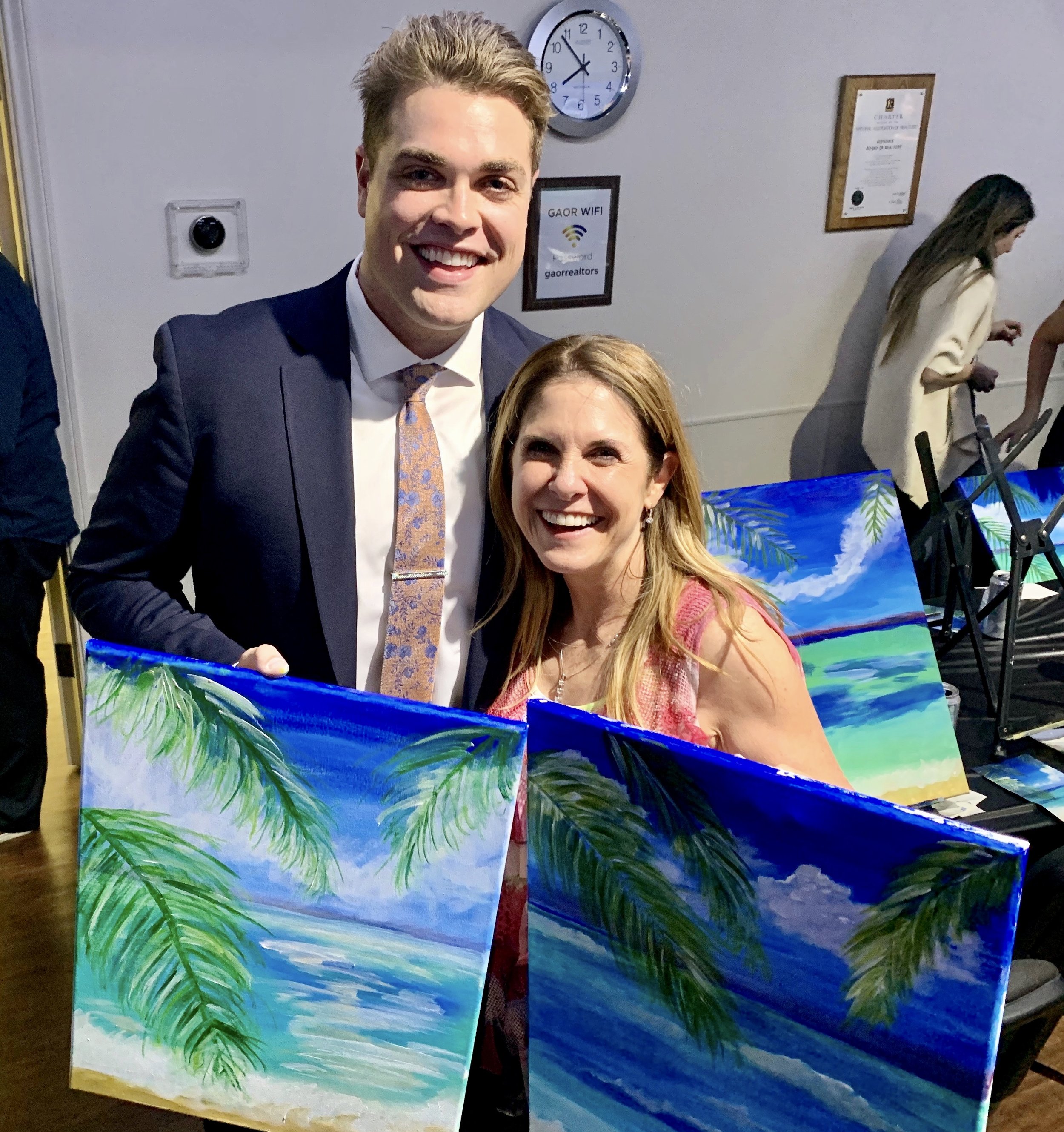 A smiling man in a suit and a woman with long hair and a pink top, both standing behind colorful beach scene paintings on canvases, at an indoor event.