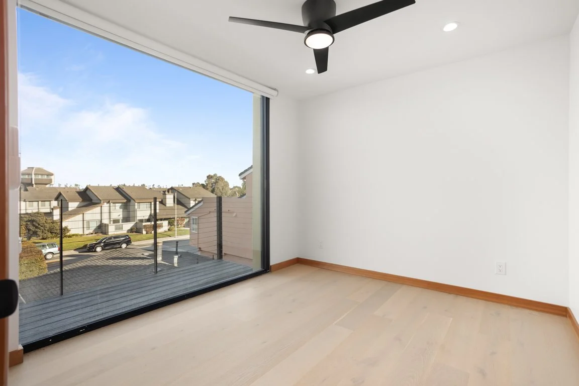 Empty room with large sliding glass door opening to a balcony, white walls, light wood floor, black ceiling fan, and recessed lighting.