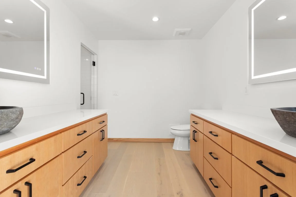 Modern bathroom with white walls, wooden cabinets, large mirrors with LED lights, a toilet in the back, and stone sinks on the counters.