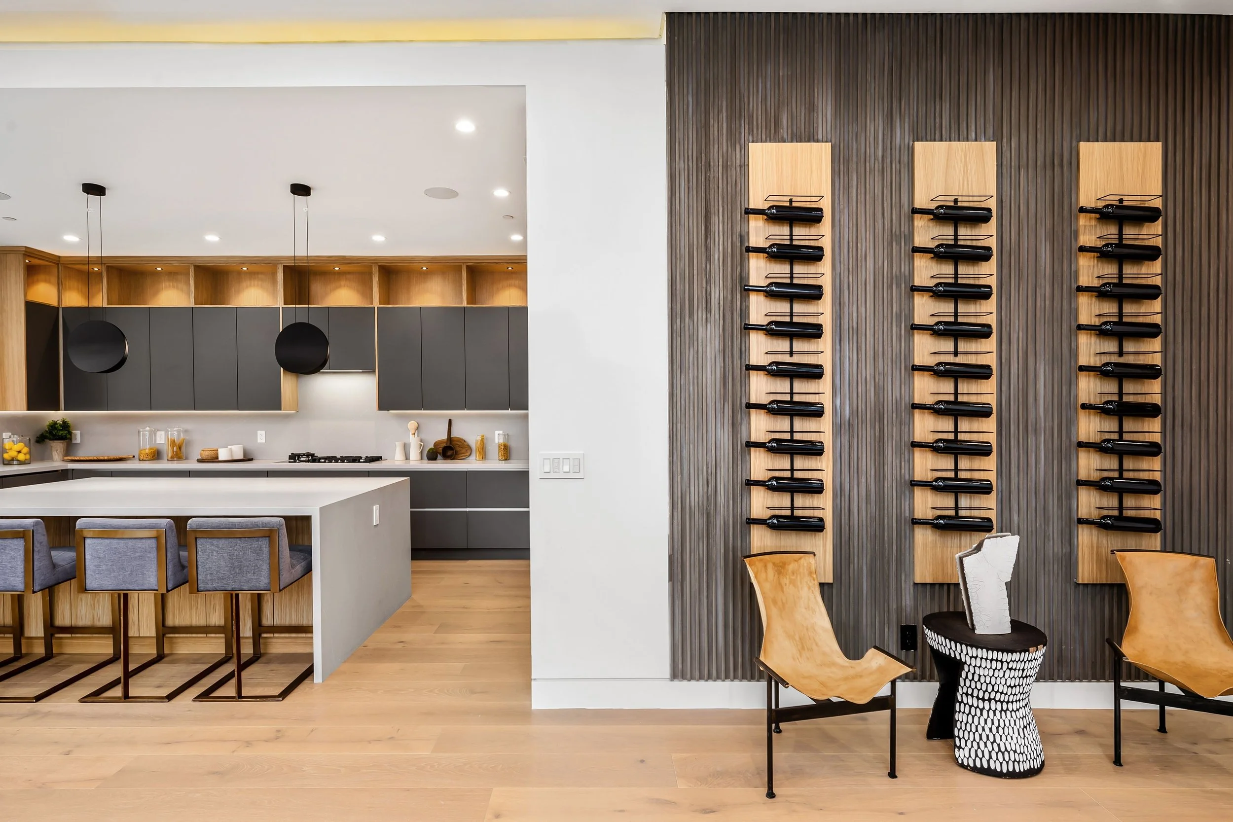 Modern kitchen and wine storage area with wooden and gray cabinetry, bar stools at a white island, and wall-mounted wine racks with bottles.