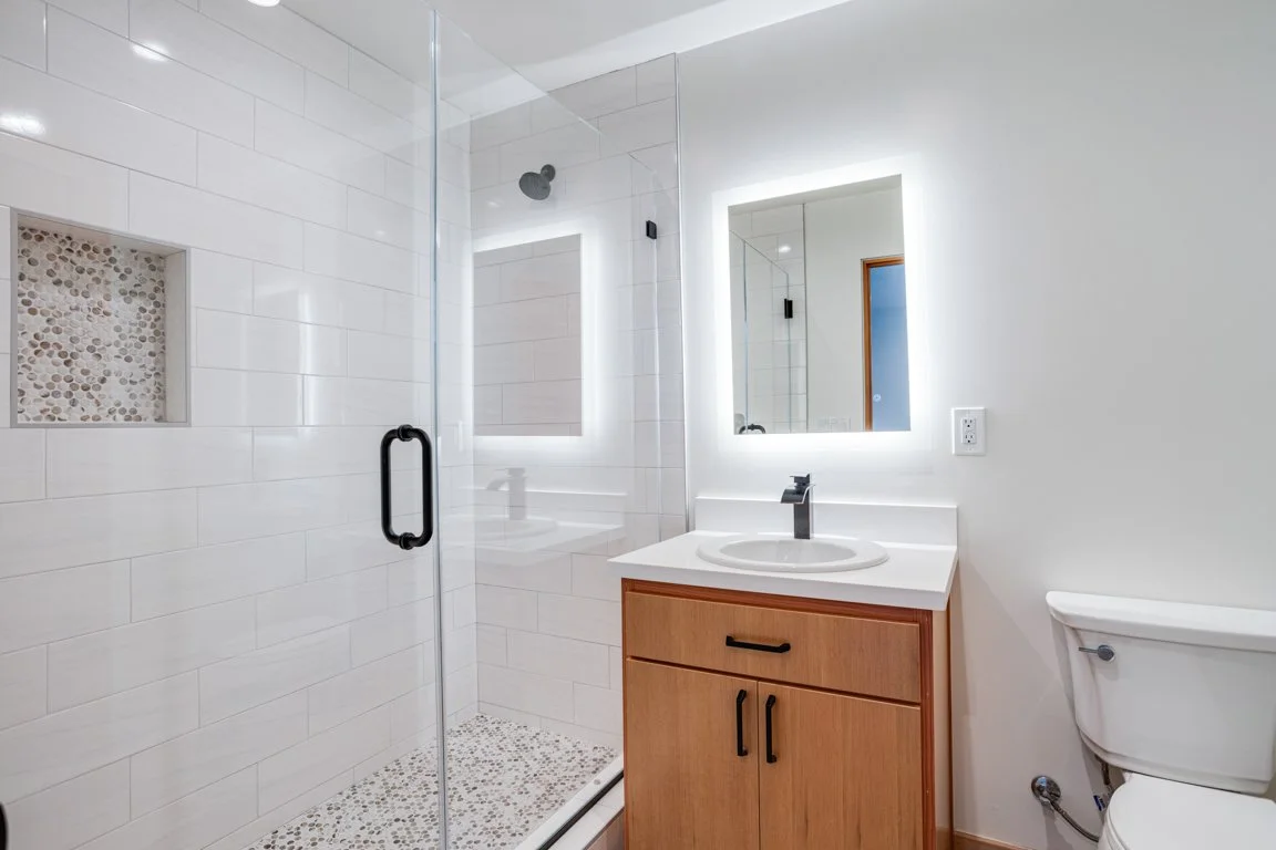Modern bathroom with a walk-in shower featuring a pebble tile floor, white tiled walls, a wall-mounted showerhead, a wooden vanity with a white sink, a illuminated mirror, and a toilet.