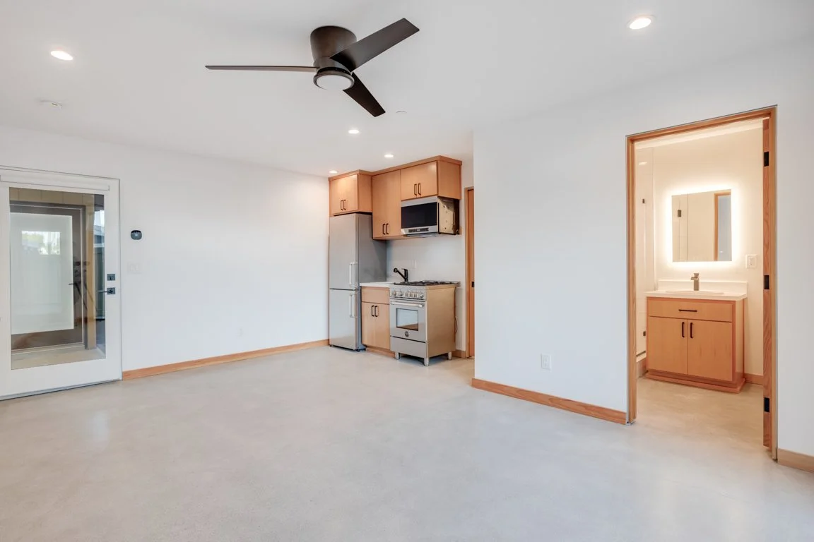 Empty living room with a ceiling fan, kitchen area with appliances, and a separate bathroom or vanity area with a mirror and sink.