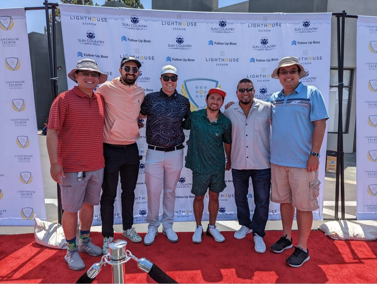 Group of six men standing on a red carpet in front of a step and repeat banner, smiling with arms around each other, at a Lighthouse Escrow event. They are wearing casual summer clothing and hats, with sunglasses.