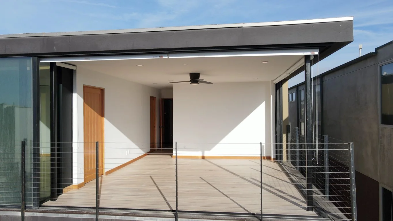 Empty outdoor balcony with wooden flooring, black metal railing, white walls, a black ceiling fan, and sliding glass doors, attached to a modern apartment building.