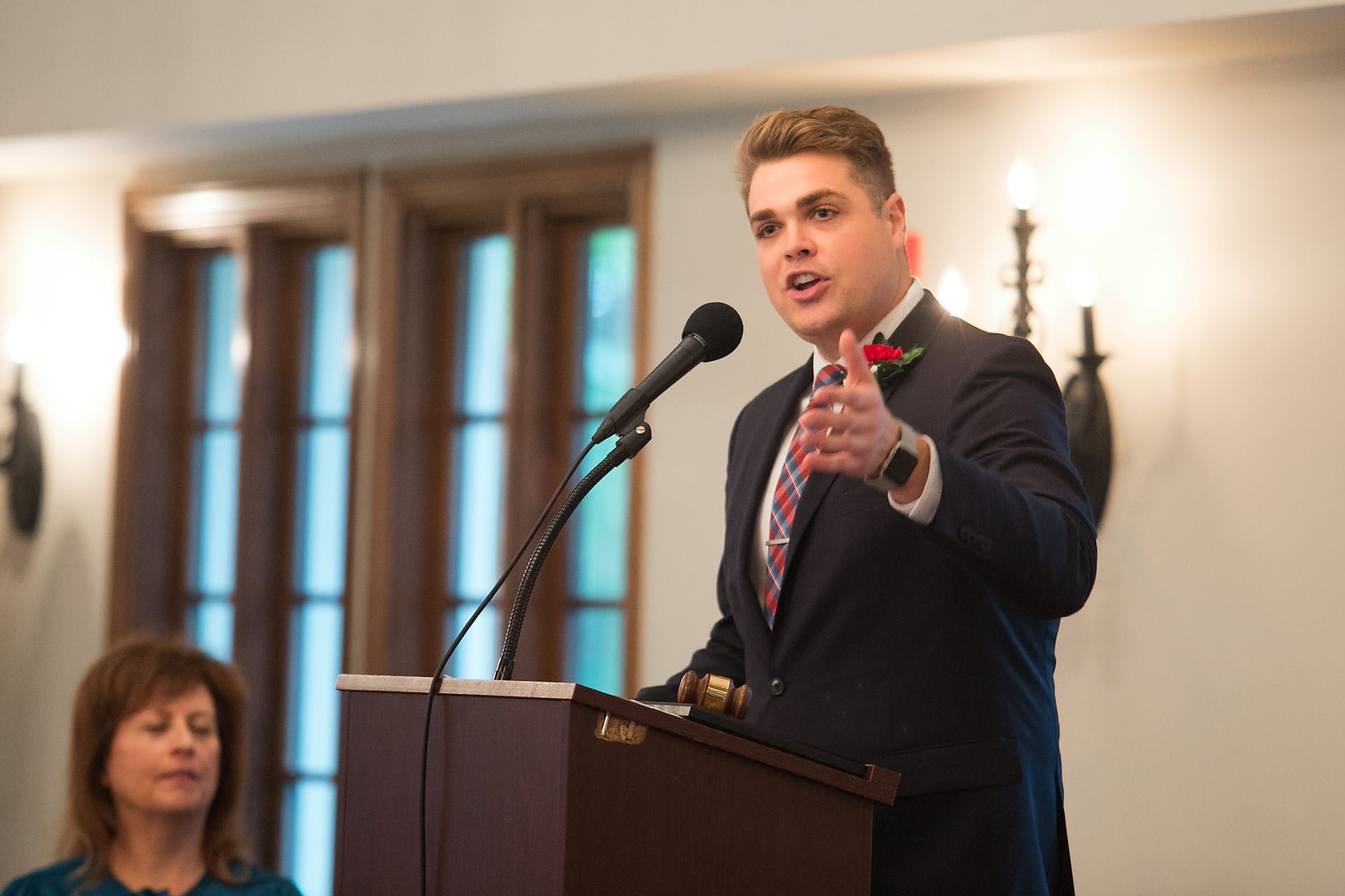 A young man in a suit giving a speech at a podium with a gavel, gesturing with his right hand, and a woman in the background listening.