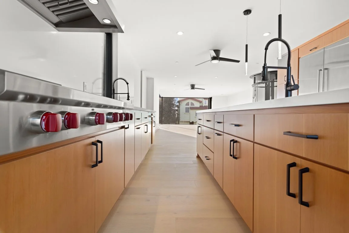 Modern kitchen with light wood cabinets, a large window, black faucet, and a stainless steel gas stove with red control knobs.