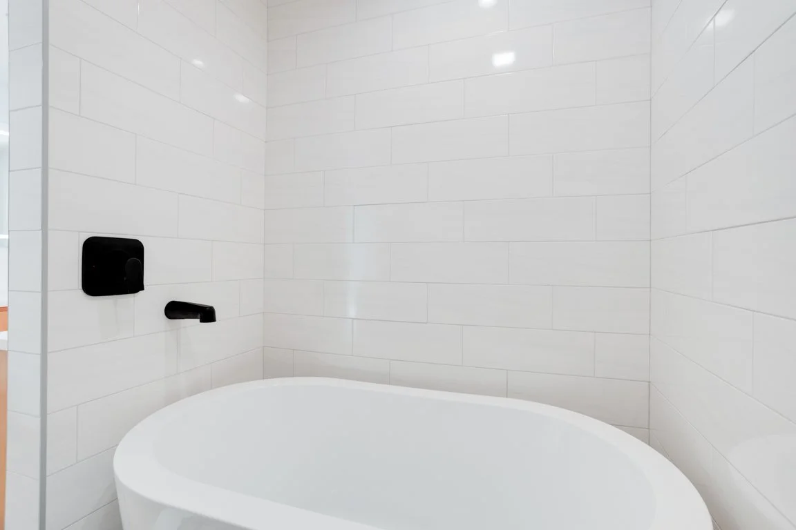Empty bathroom corner with white tiled walls and a white bathtub, black wall-mounted faucet, and a black soap dispenser.