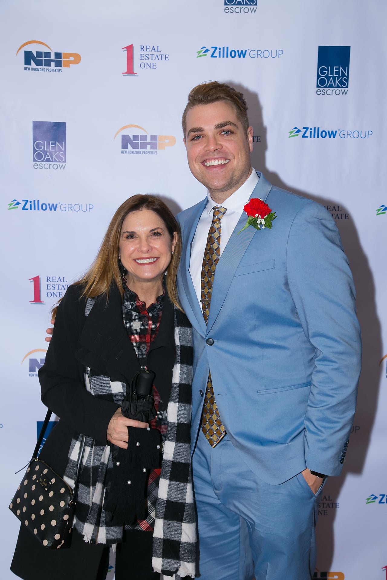 A woman and a man dressed in a light blue suit and a checkered tie are smiling and posing for a photo at a real estate event. The woman is holding a camera with a black and white checkered scarf, and the man has a red flower pinned to his lapel. The 